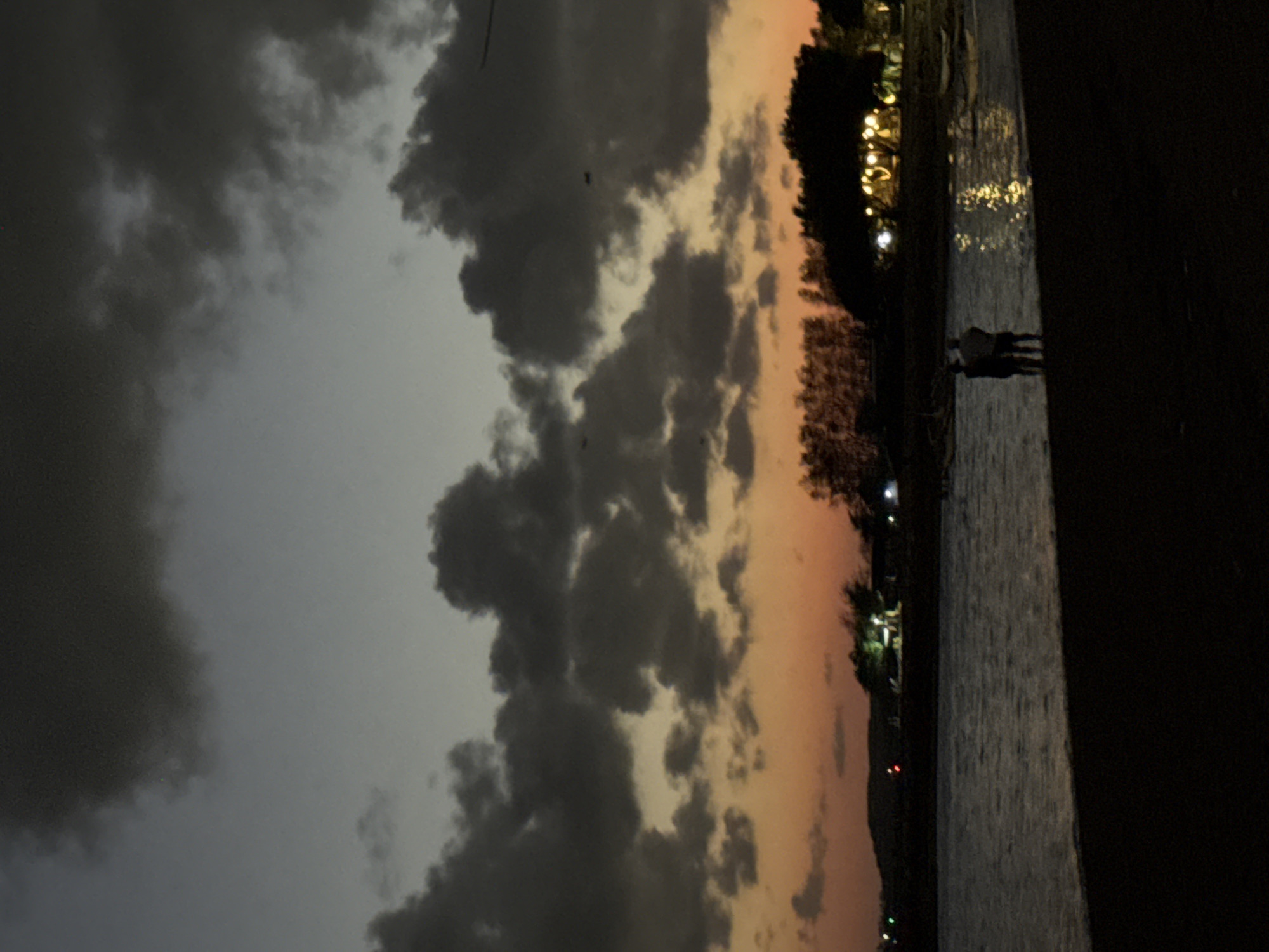 The image depicts a twilight scene at a beach or waterfront, likely in Denpasar, Indonesia, given the context provided. The sky is a gradient of deep gray clouds and a warm orange hue near the horizon, suggesting the sun has recently set or is about to rise. Dark, heavy clouds dominate the upper portion of the frame. Below the sky, silhouetted trees and structures are visible along the distant shore, with some illuminated lights suggesting human activity or dwellings. The water in the foreground is calm, reflecting the lights from the shore. A dark foreground suggests a beach or embankment. Two indistinct figures, appearing to be a couple, stand at the edge of the water, facing away from the camera and towards the illuminated shore. Their posture suggests they are observing the scenery or each other. The overall atmosphere is serene and contemplative, characterized by the muted colors and the quiet presence of the two figures.