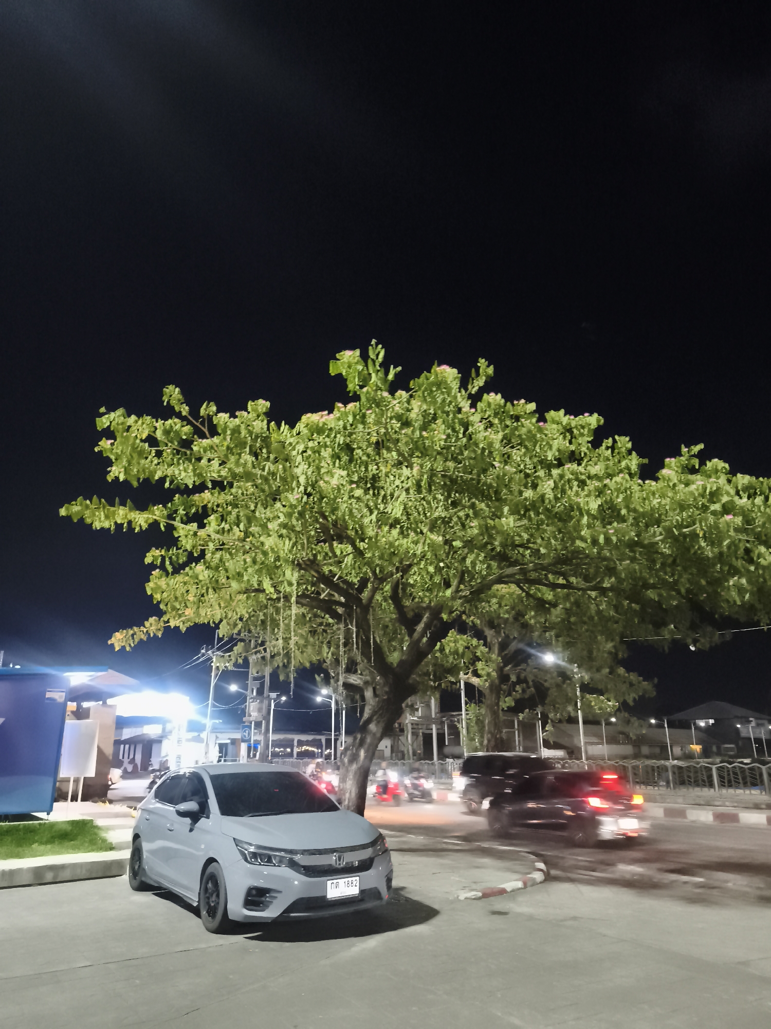 A light-gray Honda City hatchback is parked on the side of the road. Behind the car is a large tree. In the background, blurred lights from passing vehicles and buildings are visible. The license plate of the parked car is visible. The location is Baan Chaweng, Thailand. The time is night. The sky is dark.