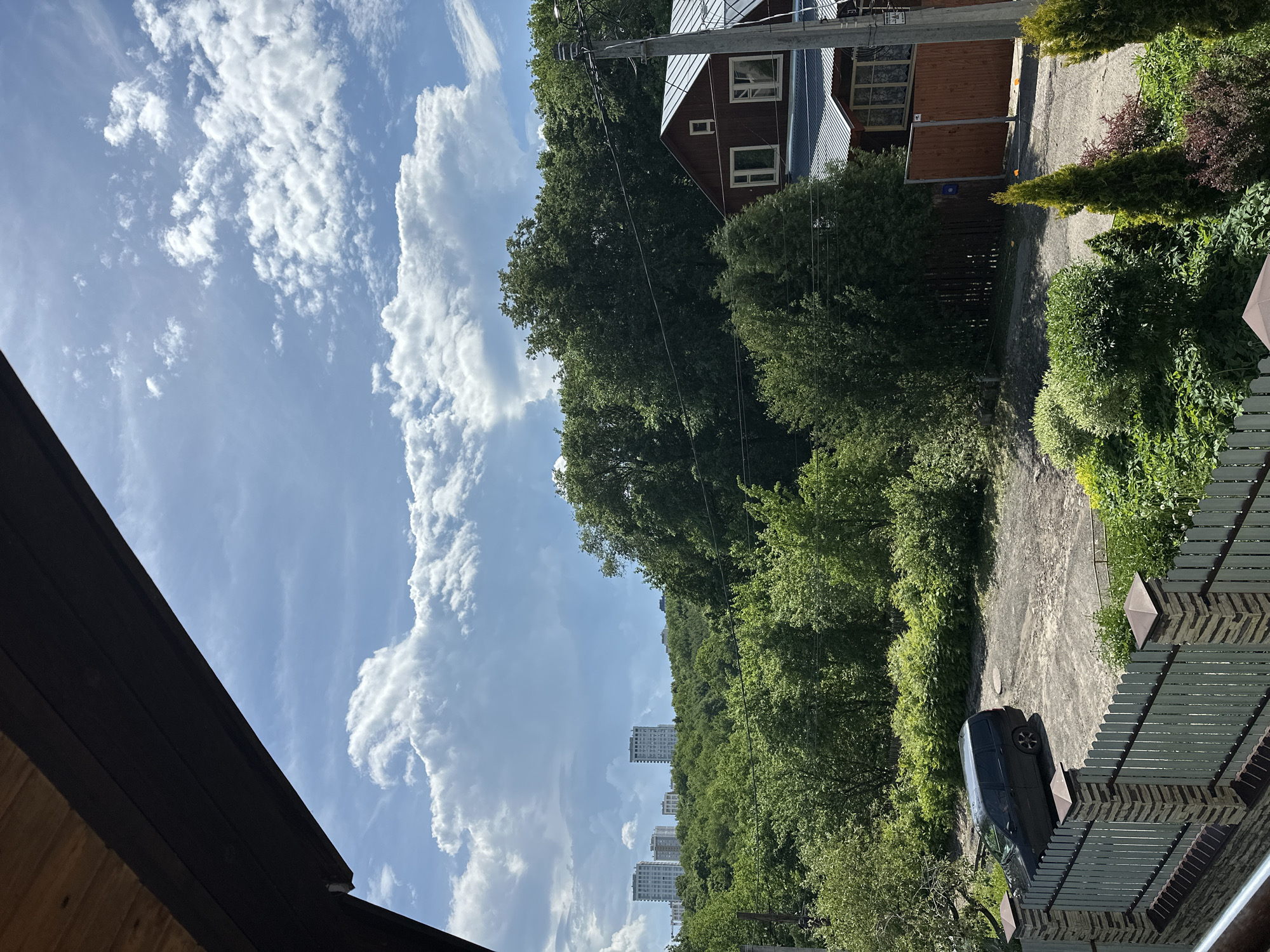 The image depicts an outdoor scene, likely a residential area, in Nizhny Novgorod, Russia. The sky occupies a significant portion of the frame, displaying a mix of light blue and cumulus clouds. The foreground includes portions of rooftops from nearby structures.
In the middle ground, a dense array of trees and foliage is visible. Apartment buildings can be seen peeking above the trees, suggesting the scene is in an urban or suburban area. A road or driveway is also present. A dark-colored minivan is parked on the driveway, angled toward the left.
Elements of residential infrastructure are scattered throughout the image, including fences and power lines. The environment seems lush and green, typical of a summer scene in this geographic location.