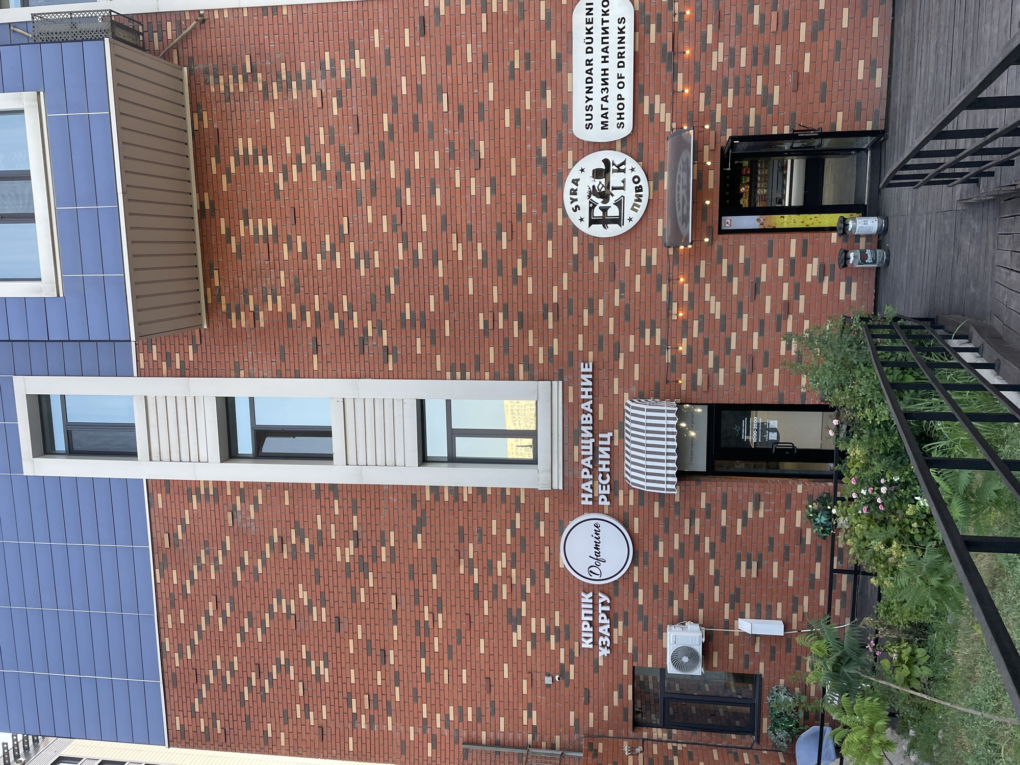 The image shows the exterior of a building with a brick facade, featuring multiple signs for businesses. On the left, there is a sign that reads "KIPLIK UZARTY" and another with a circular logo that says "Dofamine." Next to these is a sign for "NARASHIVANIJE RESNITC." To the right, there are two more signs: one circular with "SYRA ELK PIVO" and another rectangular one that states "SUSYNDAR DÜKENI MAGAZIN NAPITKO SHOP OF DRINKS." The building has several windows and an awning above a doorway. There are also some plants and an outdoor seating area visible. The overall impression is that of a commercial street in a city.