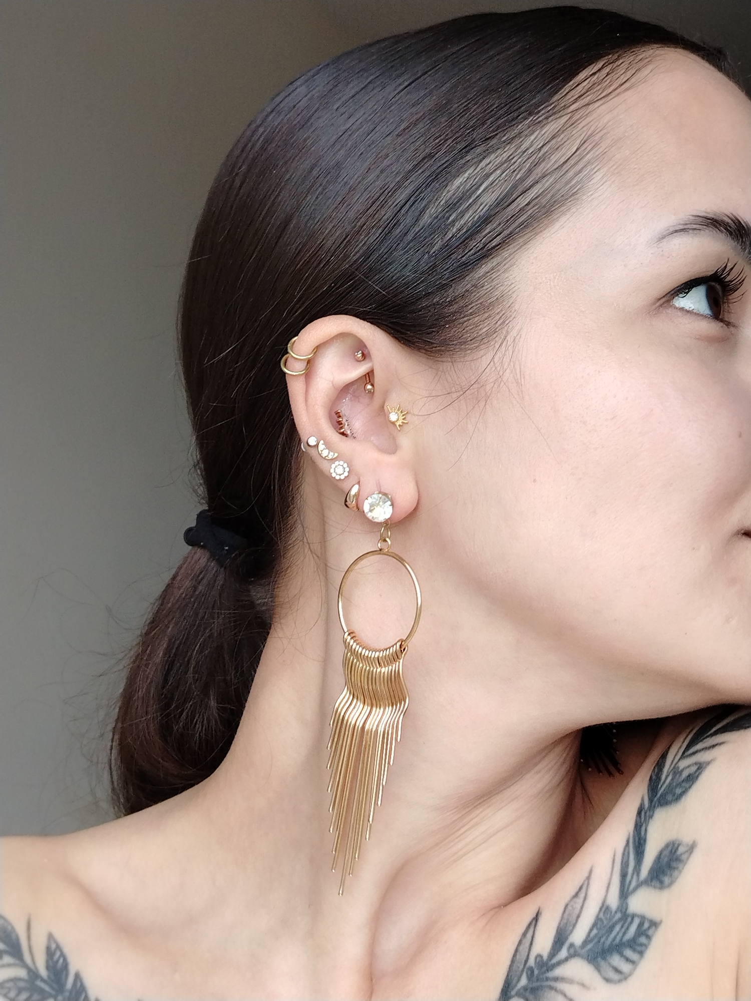A woman with dark hair pulled back into a ponytail is shown in a close-up profile shot. She is fair-skinned with visible tattoos of leafy branches on her upper chest and shoulder. Her ear is adorned with multiple gold piercings, including two rings at the top, several studs along the helix, and a long dangling gold earring with a round gem at the top, consisting of a large hoop with multiple gold strands hanging from it. She is looking slightly upward and to the right. The background is a plain, neutral gray. There is no evidence of the city of Noviy, Russia, or any specific location.