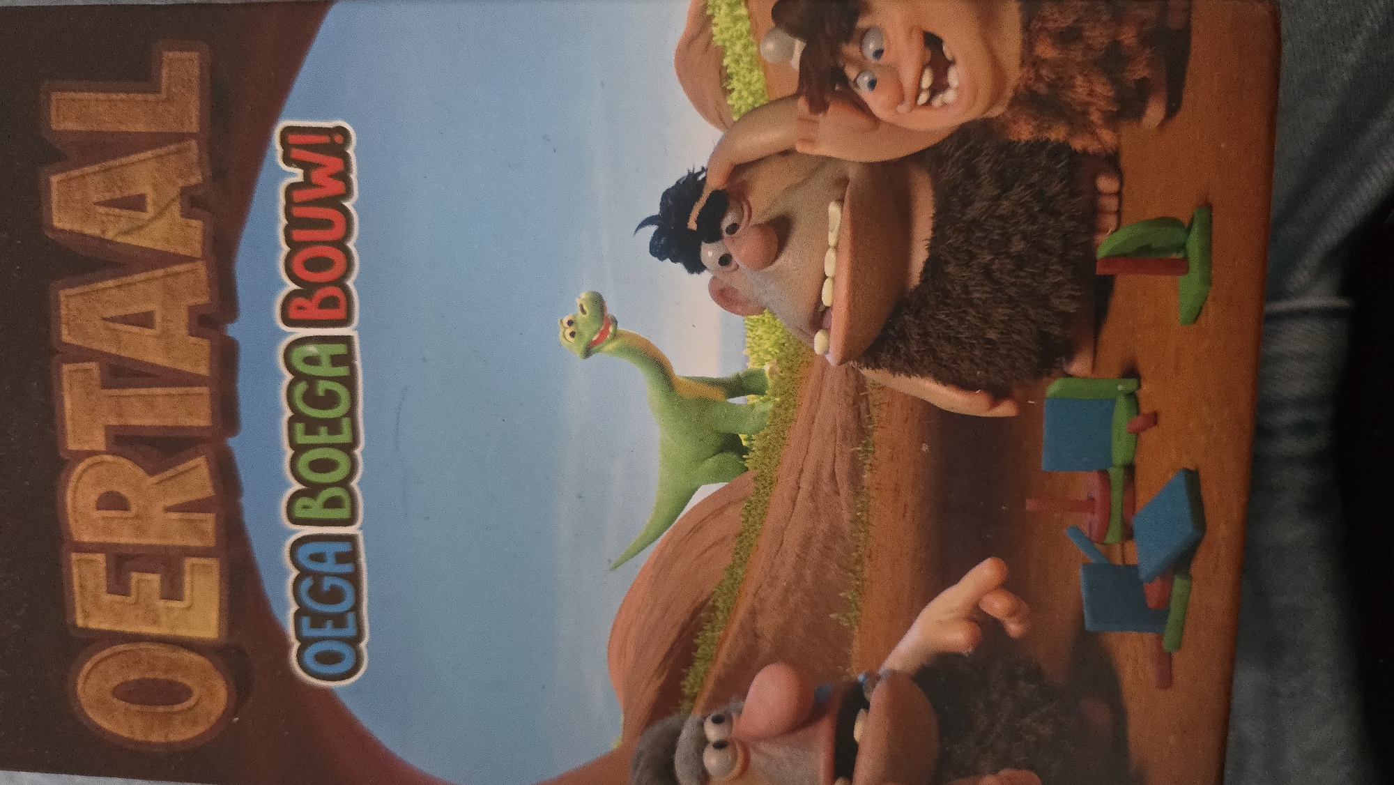 The image shows the DVD cover of a children's film featuring cartoon caveman characters and a green dinosaur against a blue sky and brown terrain backdrop. The title "OERTAAL" is printed vertically in brown letters on the left, with the phrase "OEGA BOEGA BOUW!" next to it in smaller, colorful lettering. To the right of the title are the cartoon characters and dinosaur. Below and in front of the cavemen are blue and green toy blocks and pegs. The photograph appears to be taken indoors, possibly on a table.
