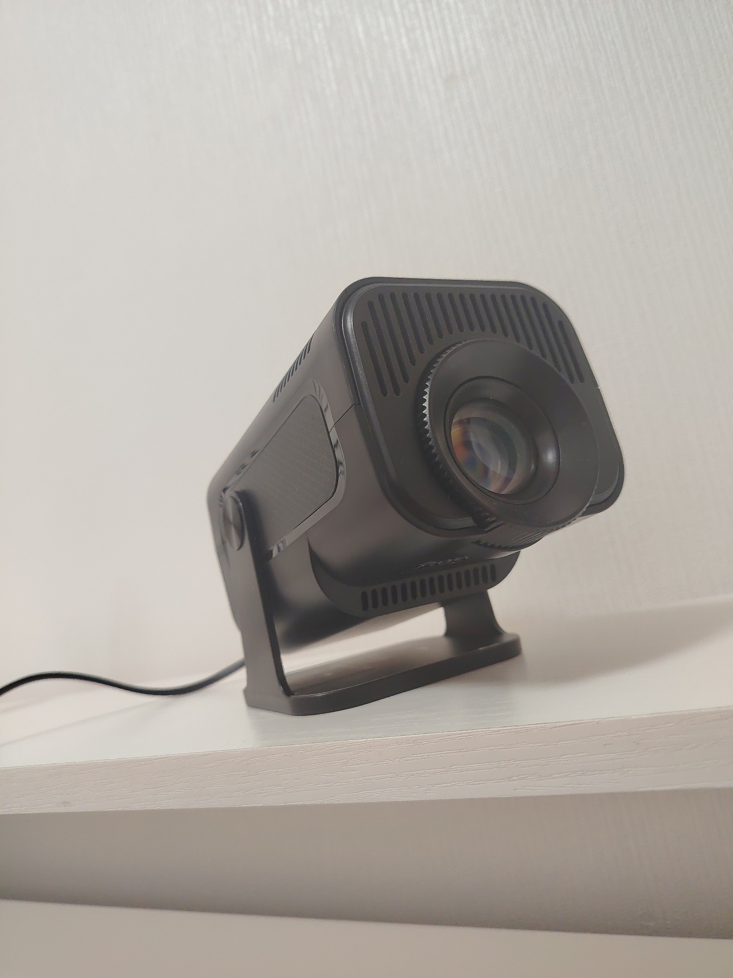 The scene, located indoors in Yekaterinburg, Russia, features a dark gray or black compact digital projector positioned on a light-colored, horizontally textured shelf. The projector is angled slightly to the viewer's right and upward. It exhibits a prominent, multi-element projection lens on its front face, framed by a serrated focus ring. Vertical ventilation grilles are visible on the top and sides of the device, along with a textured, possibly speaker, panel on its left side. The projector rests on a dark, U-shaped support stand. A black power cable extends from the rear of the unit, trailing across the light-colored shelf. The background consists of a plain, off-white wall with subtle, vertically oriented striations, suggesting a fine-textured finish.