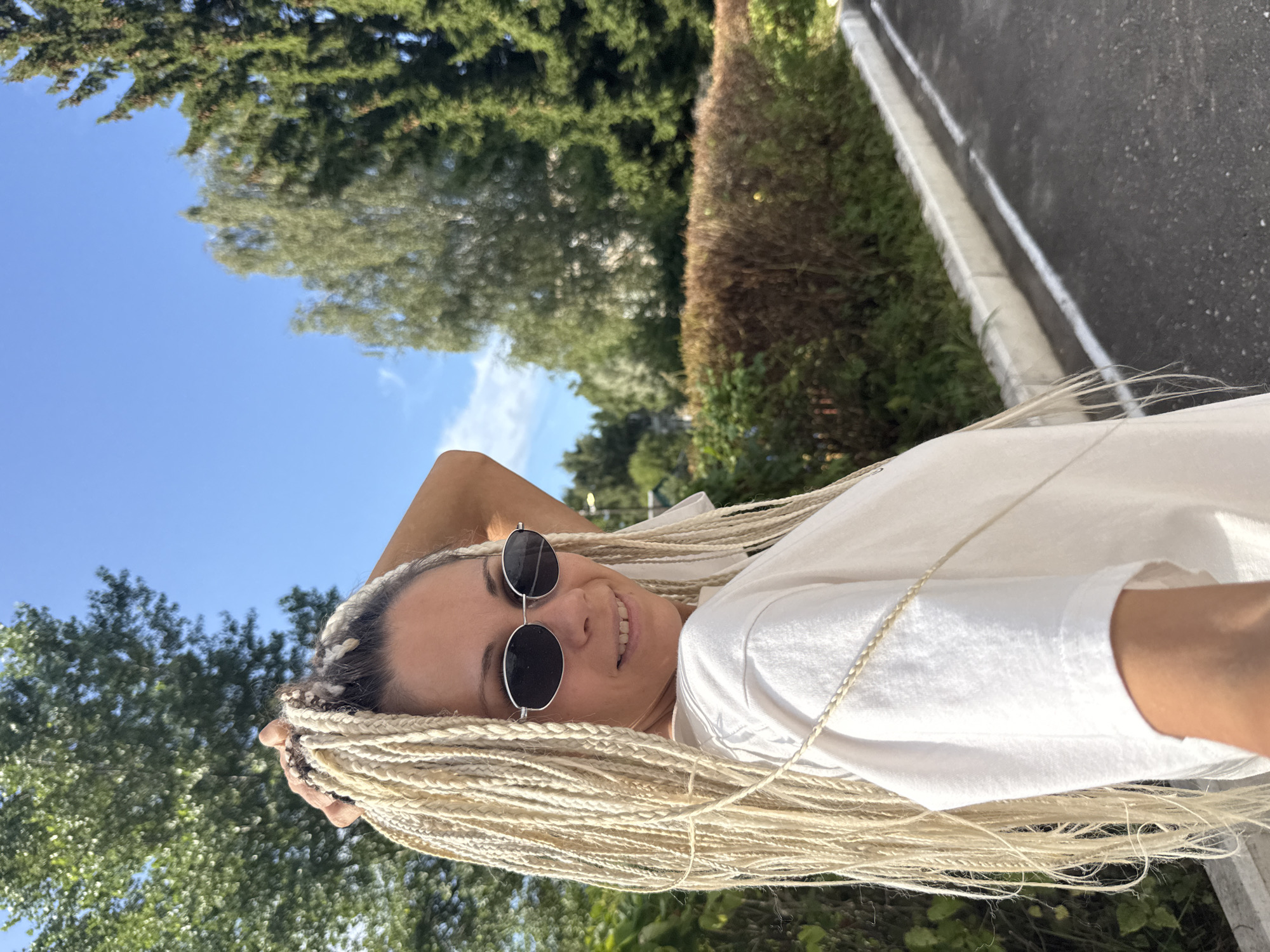 In Rusanovo, Russia, a woman with blonde braided hair and sunglasses is pictured outdoors. She is wearing a white t-shirt and her right hand is raised, holding her hair. The background features green trees, a brown hedge, a road with a white line, and a blue sky with some clouds.