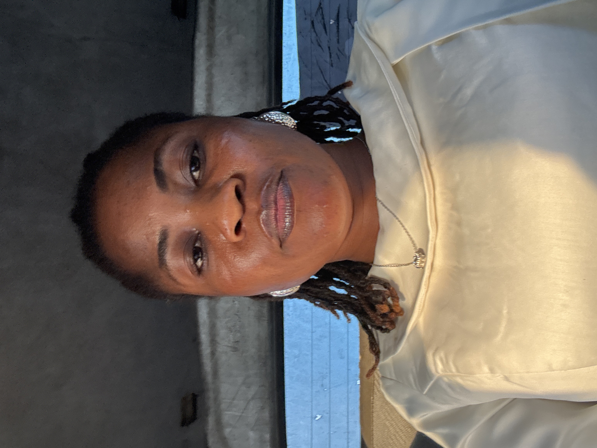 A frontal medium shot shows a dark-skinned woman with braided hair, wearing a cream-colored top and earrings, and a necklace. She is looking at the camera. The background appears to be the interior of a vehicle, with a grey headliner and a blue window. The image is taken in Ajara, Nigeria.