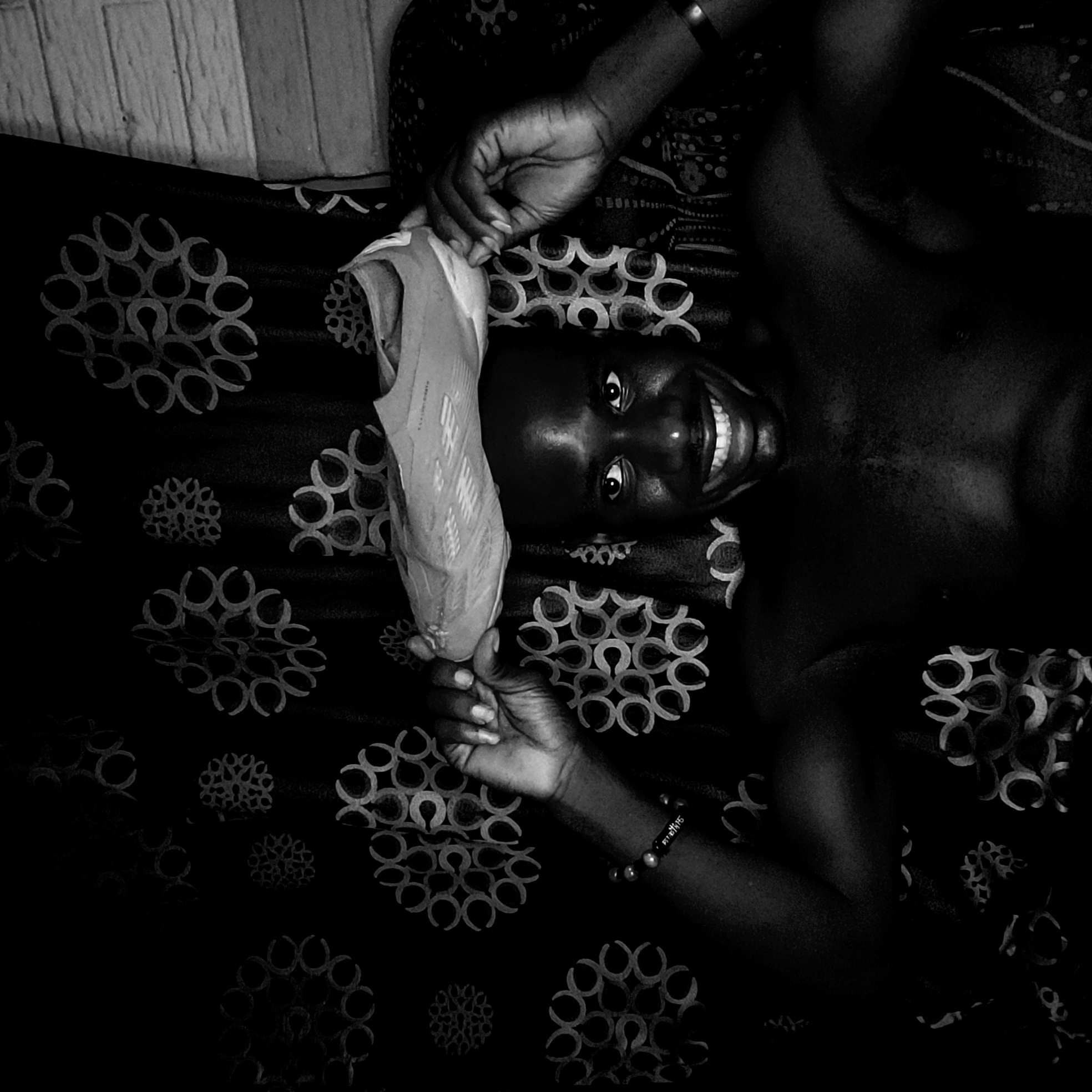 A black and white, high-angle portrait features an individual with dark skin lying on their back, smiling broadly, and holding a light-colored sports shoe. The person is looking directly at the camera with wide, joyful eyes and a prominent, toothy grin, conveying happiness and engagement.
Their upper torso and arms are visible, resting on a dark patterned fabric, possibly a bedspread or blanket, adorned with circular and floral-like designs. A dark bracelet is visible on their right wrist. The light-colored shoe, held with both hands near the person's head, appears to be an Adidas soccer cleat. Visible text on the shoe includes "ADIDAS" and elements that suggest "X SPEEDFLOW" or "X SPEEDPORTAL," although these are partially obscured. In the top left corner of the frame, a light-toned wooden-paneled surface, possibly a wall or headboard, can be seen.
The scene is set indoors, likely a bedroom. The monochromatic tone of the image emphasizes textures and expressions. Based on the provided context, the location is Narayi, Nigeria.