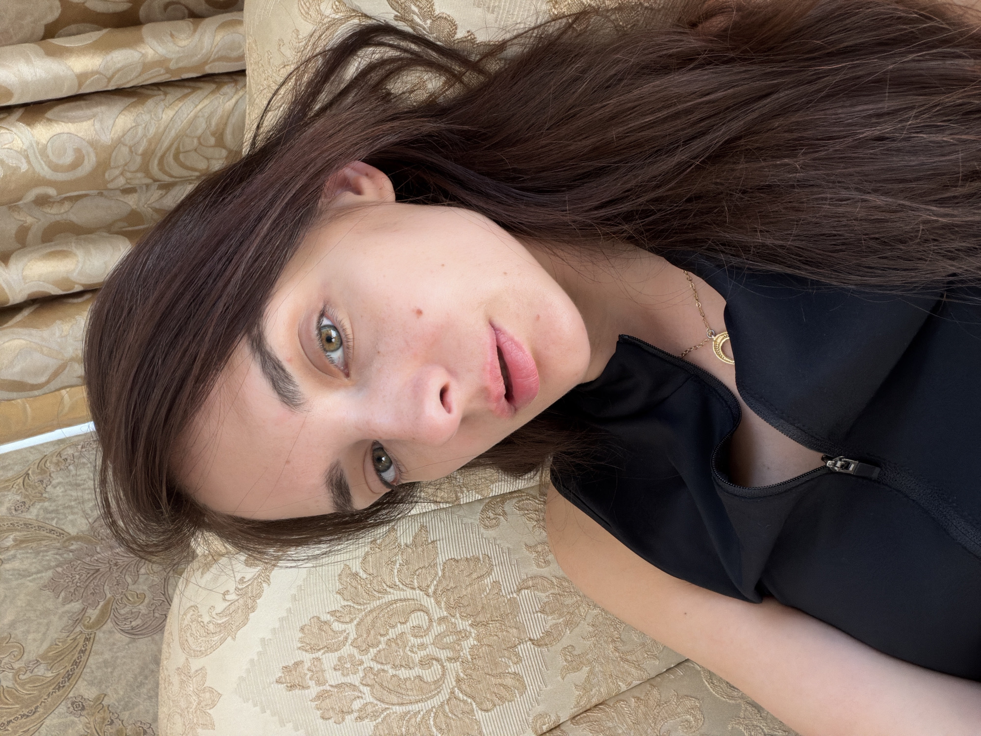 A young woman with long, dark brown hair is lying on her side on a couch. She is wearing a black, sleeveless top with a zippered neckline and a gold necklace. Her head is propped up on a patterned cushion, and she is looking directly at the camera. The background consists of patterned curtains.