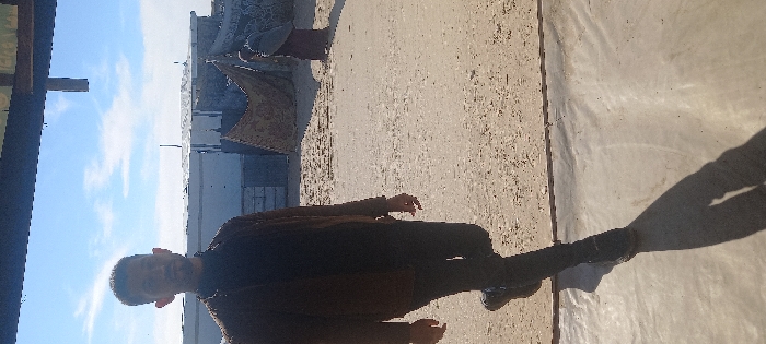 A vertical image depicts an outdoor scene in Khan Yunis, Palestinian Territory. In the foreground, a male individual, appearing in his late 20s to early 30s with short dark hair and a beard, walks rightwards, partially in shadow. He wears a dark brown jacket over a dark top, dark trousers, and dark shoes. His elongated shadow is cast on a light-colored, smooth surface in the immediate foreground. The midground is an unpaved area of light-colored sand or gravel. In the background right, a second individual, dressed in dark clothing and a head covering, is partially visible, facing away. Behind the figures, structures include a light-colored cinder block wall on the left, a draped patterned fabric or tarp, and several simple, low-rise buildings with flat roofs under a clear blue sky with scattered white clouds. A dark horizontal beam spans the top of the frame.