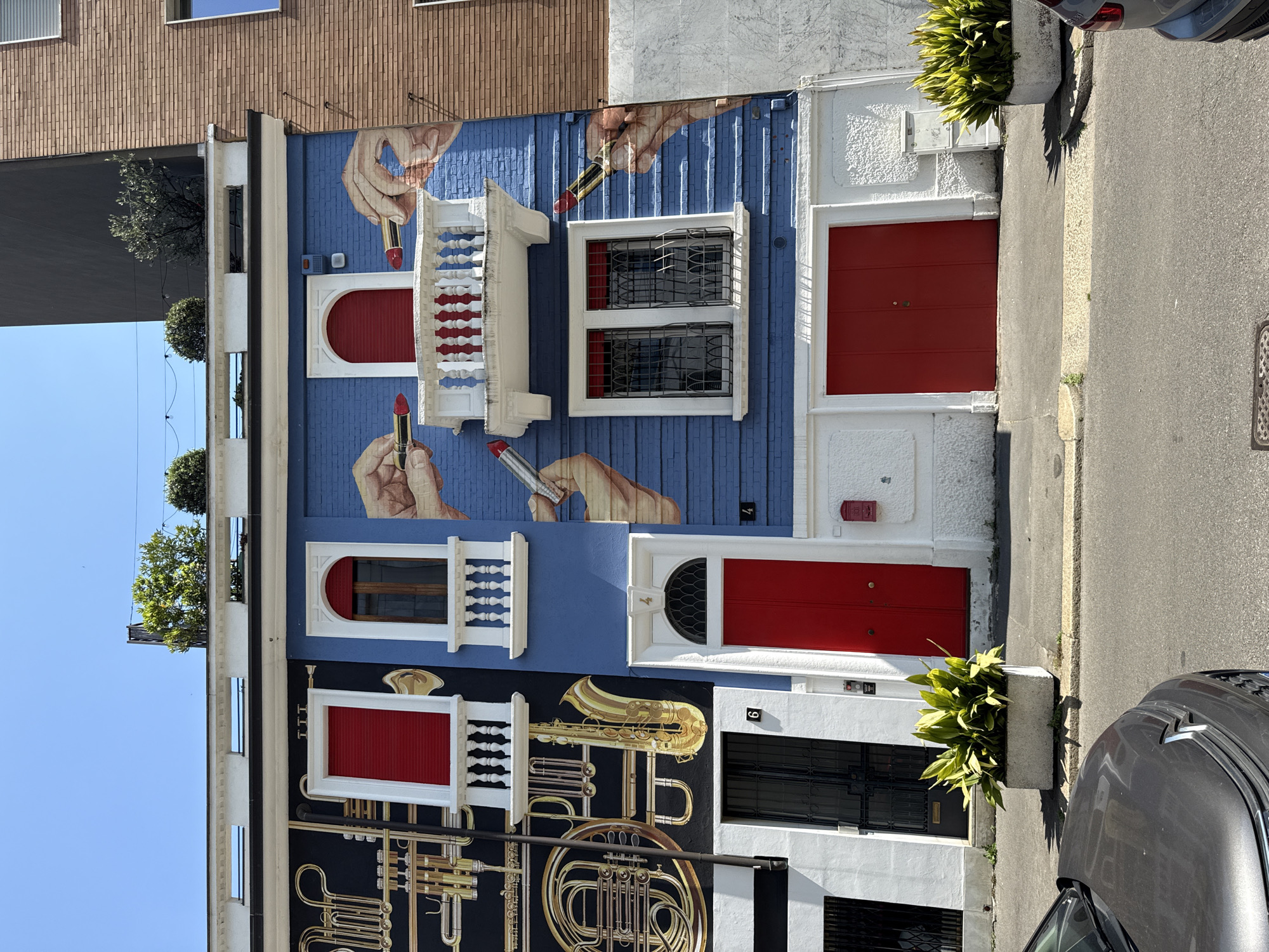 The image depicts the facades of two adjacent buildings located in the city of Milan, Italy. The first building is a three-story structure featuring a mural on its front. The mural is divided into two distinct sections. The upper section has a blue background with depictions of hands holding lipsticks. The "windows" on this section feature red blinds or are painted red. The lower section has a black background with a large mural of brass musical instruments, including a French horn and trumpets. The second building is white with black window frames. The doors of both buildings are painted red. A plant is visible in front of the second building, near the sidewalk. A portion of a parked car is visible on the right side of the image. A brick building is visible above the first building. The sky is blue and clear.