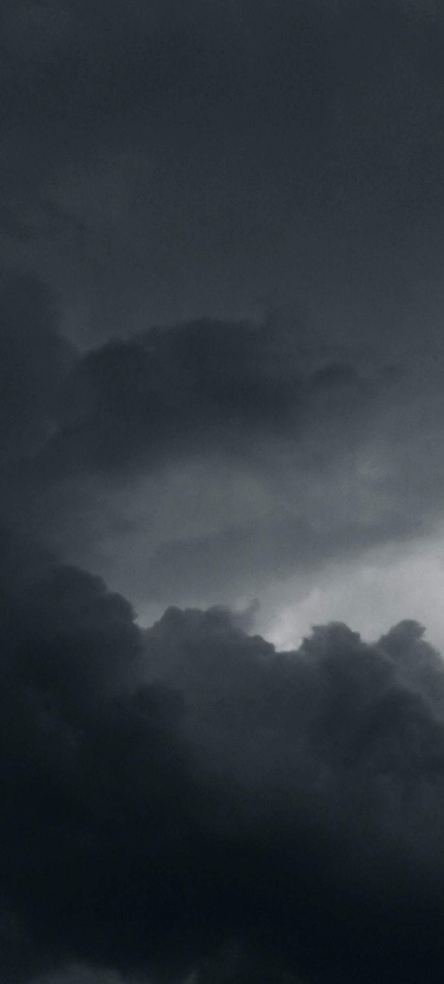 A vertical, low-angle shot captures a dramatic sky filled with dark, brooding clouds. The clouds are dense and textured, with variations in shade suggesting depth and form. Lighter, brighter areas of sky are visible through gaps in the clouds, creating contrast. The overall mood is somber and potentially stormy. The image is a cloudscape and does not contain any elements that would suggest a specific location in Winnenden, Germany, or any human presence.