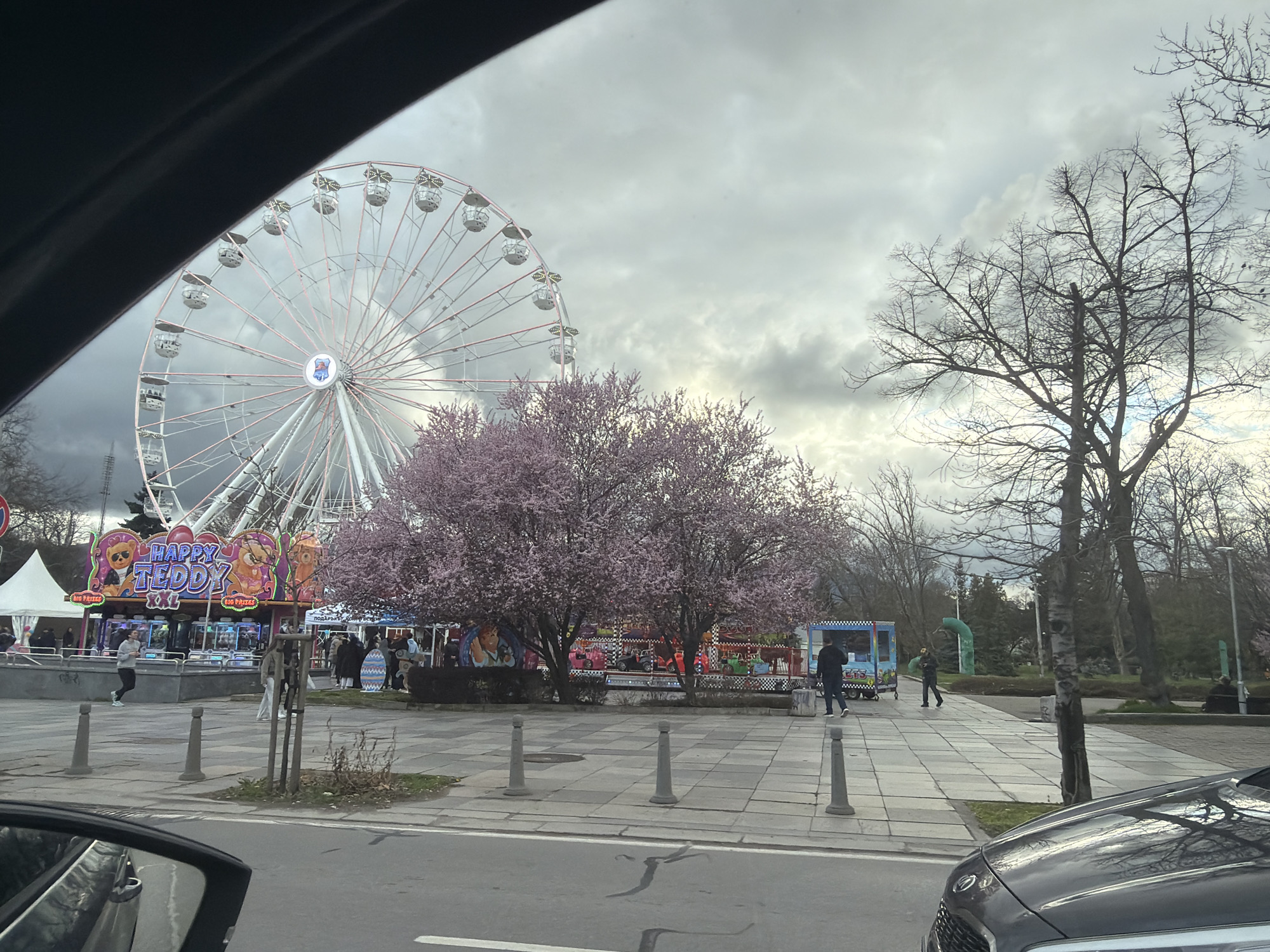 The image depicts a street scene in Sofia, Bulgaria, from the perspective of inside a car. A large Ferris wheel dominates the background. To the right of the Ferris wheel are trees, some bare and others with pink blossoms. A "Happy Teddy" prize booth, other smaller structures, and people are visible in the foreground. A pedestrian is walking on the sidewalk. Cars are parked or driving on the road. The sky is overcast.