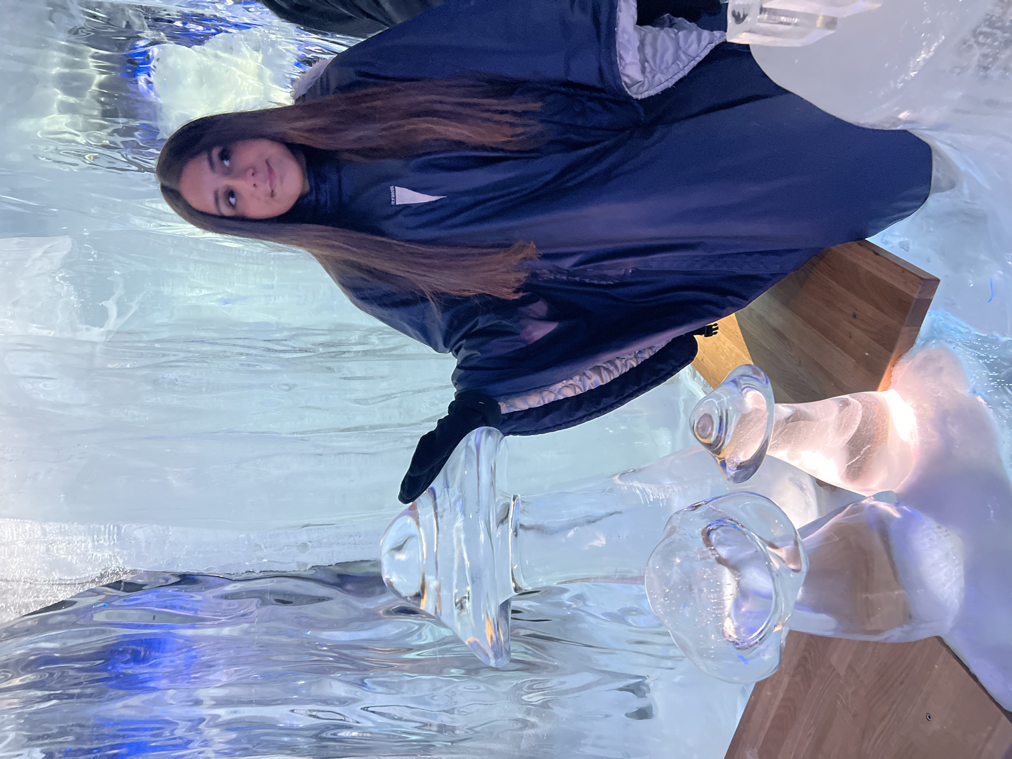 A young woman with long, dark hair stands in an ice hotel, wearing a dark blue poncho over a black turtleneck. She is wearing black gloves and is reaching out her right hand to touch a mushroom-shaped ice sculpture. The background consists of large, translucent ice walls with blue and white lighting. In the foreground, there are two more mushroom-shaped ice sculptures. One is larger and closer to the woman, while a smaller one is visible to its right. The floor is covered in a white, powdery substance that appears to be snow or ice. The scene takes place in the Icehotel, located in Jukkasjärvi, Sweden, which is a temporary hotel that is rebuilt each year using natural ice from the Torne River. It is a popular tourist attraction known for its unique ice sculptures and accommodations.