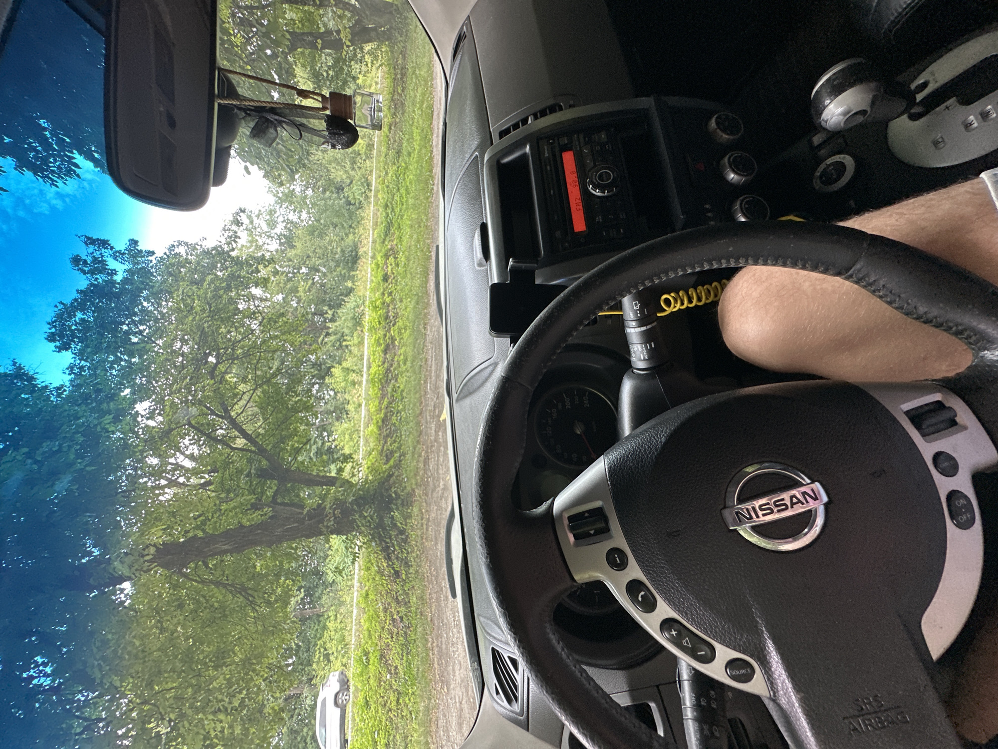 The image shows the interior of a Nissan vehicle, looking forward through the windshield. A person's leg is visible in the lower right portion of the frame. The steering wheel is in the foreground on the left, with the Nissan logo prominently displayed. The dashboard features a radio displaying "FM2 98.0", climate controls, and a gear shift. Outside the car, a tree-lined road is visible, with a white car parked on the side in the distance. The sky is blue with some clouds. The scene appears to be outdoors, likely in a rural or suburban setting, possibly in Shatava, Ukraine, given the context.