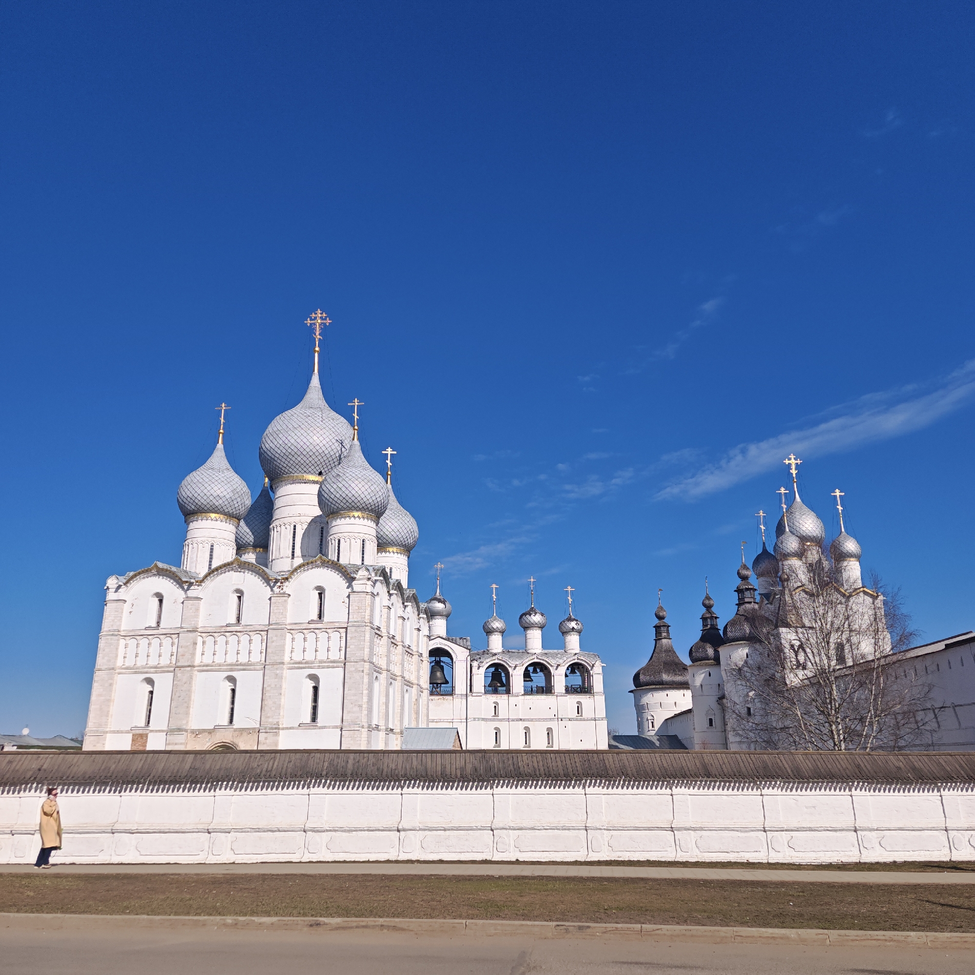 The image showcases a view of Rostov, Russia, featuring historical architecture under a clear blue sky with few clouds. On the left, a complex of buildings with silver domes topped with golden crosses rises prominently. These buildings are painted white and exhibit detailed architectural features.
A long, white wall runs horizontally across the foreground, providing a visual barrier, with a wooden section at its top. A person is seen walking alongside the wall on the left side of the frame.
In the background, behind the central complex, are additional structures with similar architectural styles and silver domes. A bare tree is visible near the complex.
The foreground also includes a strip of grass and a paved area.