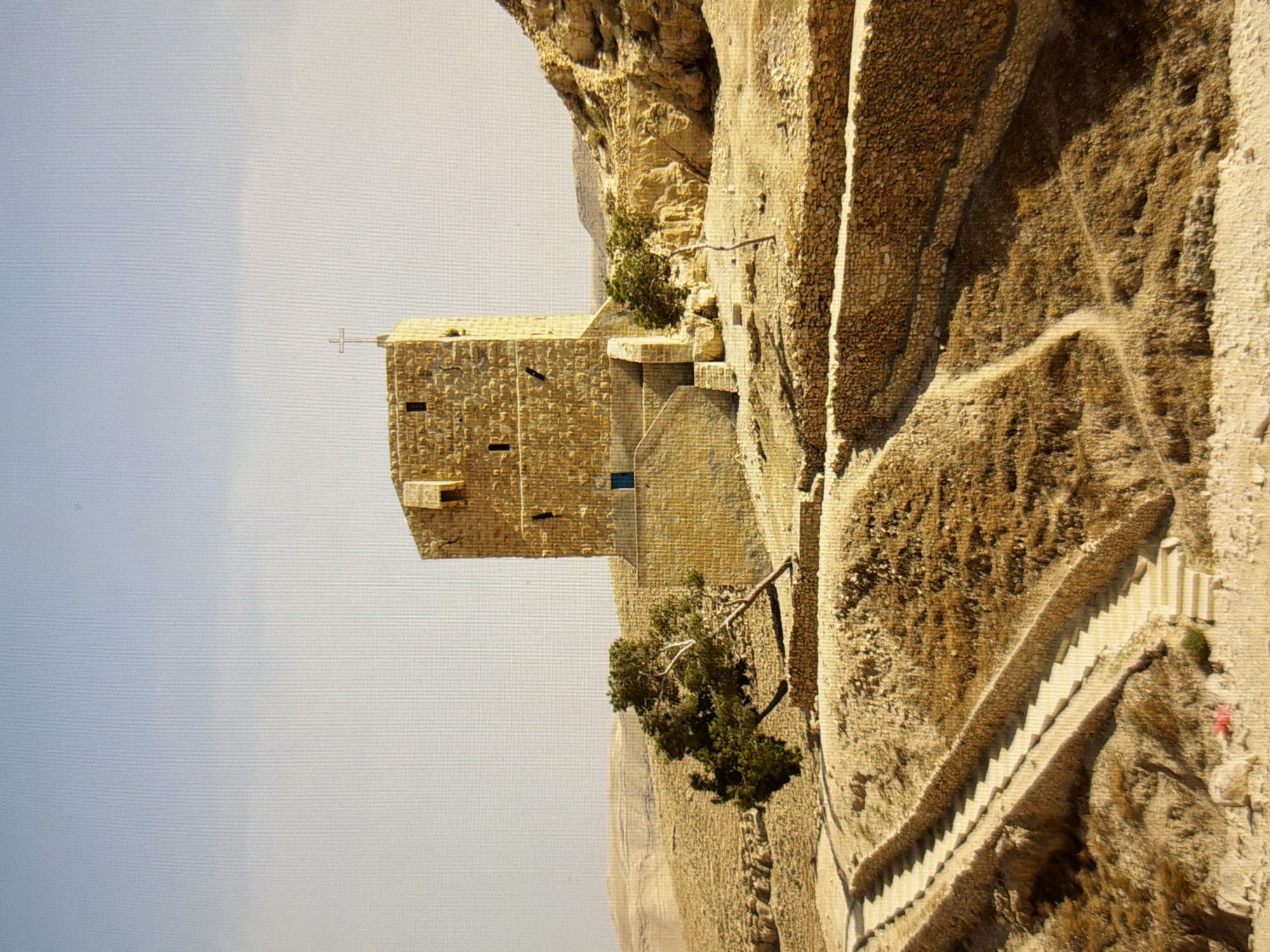 The image depicts a stone structure resembling a tower or a fortified building, situated in a dry, arid landscape with rolling hills. A cross is visible on the top of the structure. A tree with gnarled branches leans to the left in front of the building. Stone walls and pathways with stairs are present in the foreground, built into the terrain. The sky is a pale blue. There are no people visible in the image. The scene appears to be in a remote, possibly historical or religious site, characterized by its natural environment and architectural elements. The location of this image is not in the city of Kyiv, Ukraine.