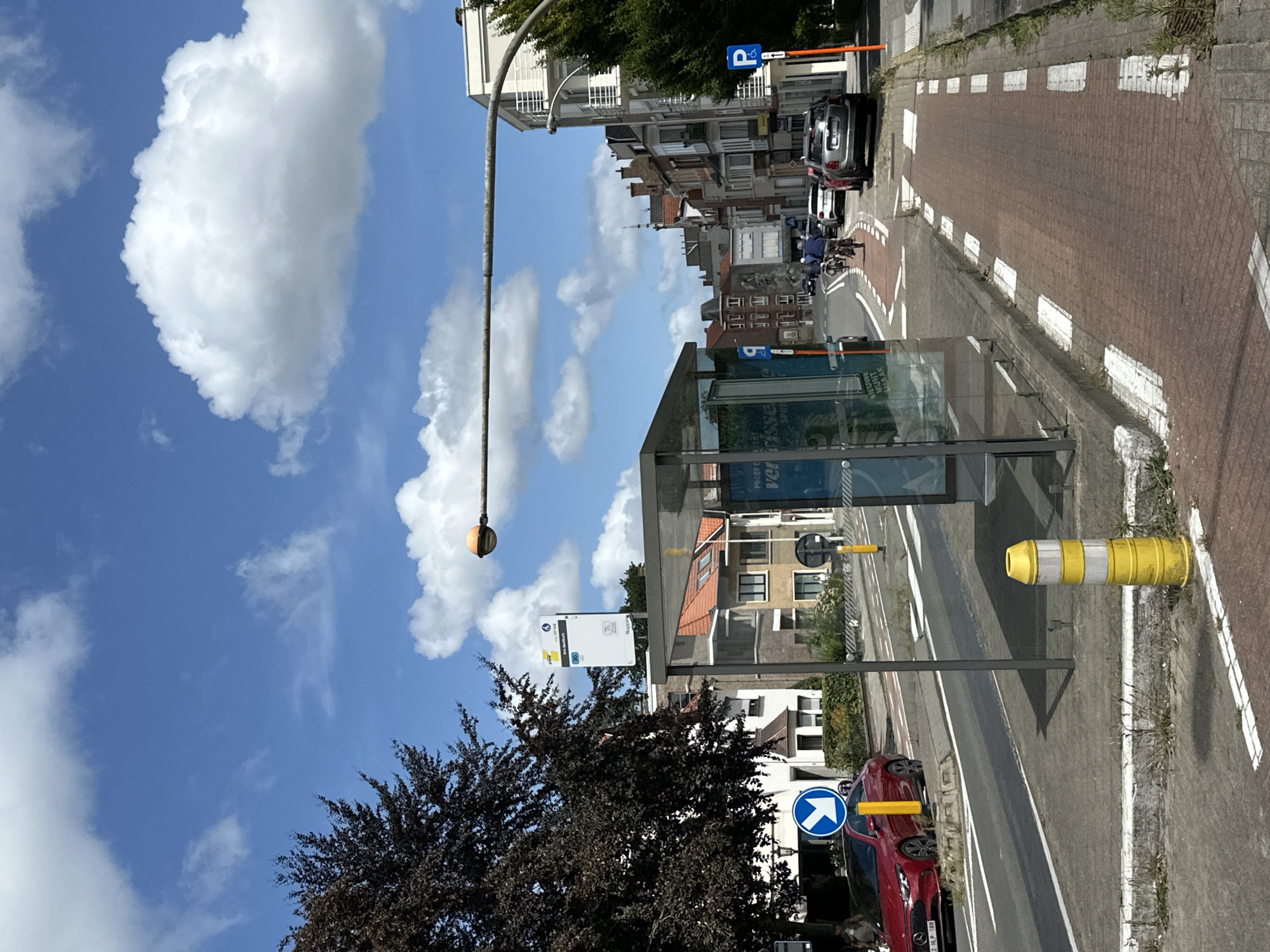 The image depicts a street scene in Bruges, Belgium, on a clear day with a bright blue sky and scattered white clouds. In the foreground, a red sedan with license plate "2-HLP-188" is parked on the left side of the road. Adjacent to the car is a bus stop shelter with glass panels. A tall, striped yellow and white bollard stands near the shelter.
The street features a designated red brick bike lane running along the right side, marked with white dashed lines and rectangular white patches. Several cyclists are visible on the bike lane in the middle ground, with one in the foreground wearing a blue shirt and another in a grey top. Beyond the cyclists, a silver-grey hatchback is parked on the right side of the street.
Buildings line the street, displaying typical European architecture with multi-story structures, red-tiled roofs, and windows. A prominent bus stop sign with route information is mounted on a pole near the shelter. A street lamp with an orange globe extends from a pole over the road. Green trees, including a dark-leaved tree on the left, add to the urban landscape. The overall environment suggests a well-maintained urban area with integrated public transport and cycling infrastructure.