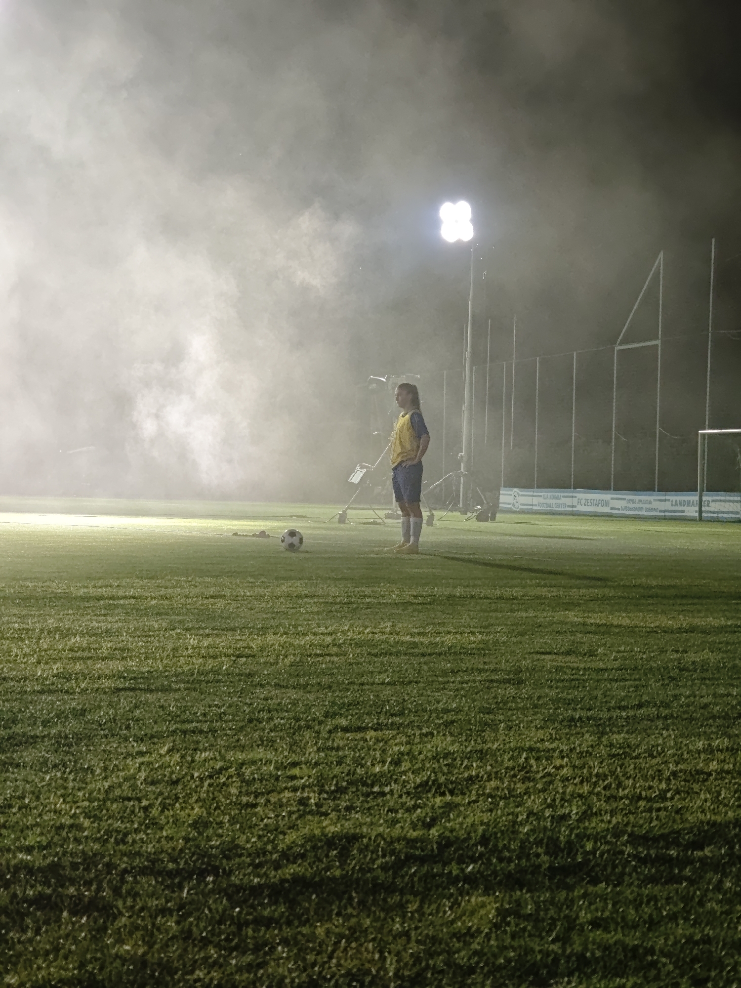 A person stands on a soccer field at night, illuminated by floodlights amidst a dense fog or smoke. The person is wearing a yellow vest over a blue and white uniform, with white socks and yellow cleats. A soccer ball is visible on the grass in the foreground to the left of the person. Behind the person, a fence and stadium lighting are discernible. The background suggests a sports facility. The location is identified as the FC Zestafoni Football Center in Tbilisi, Georgia.