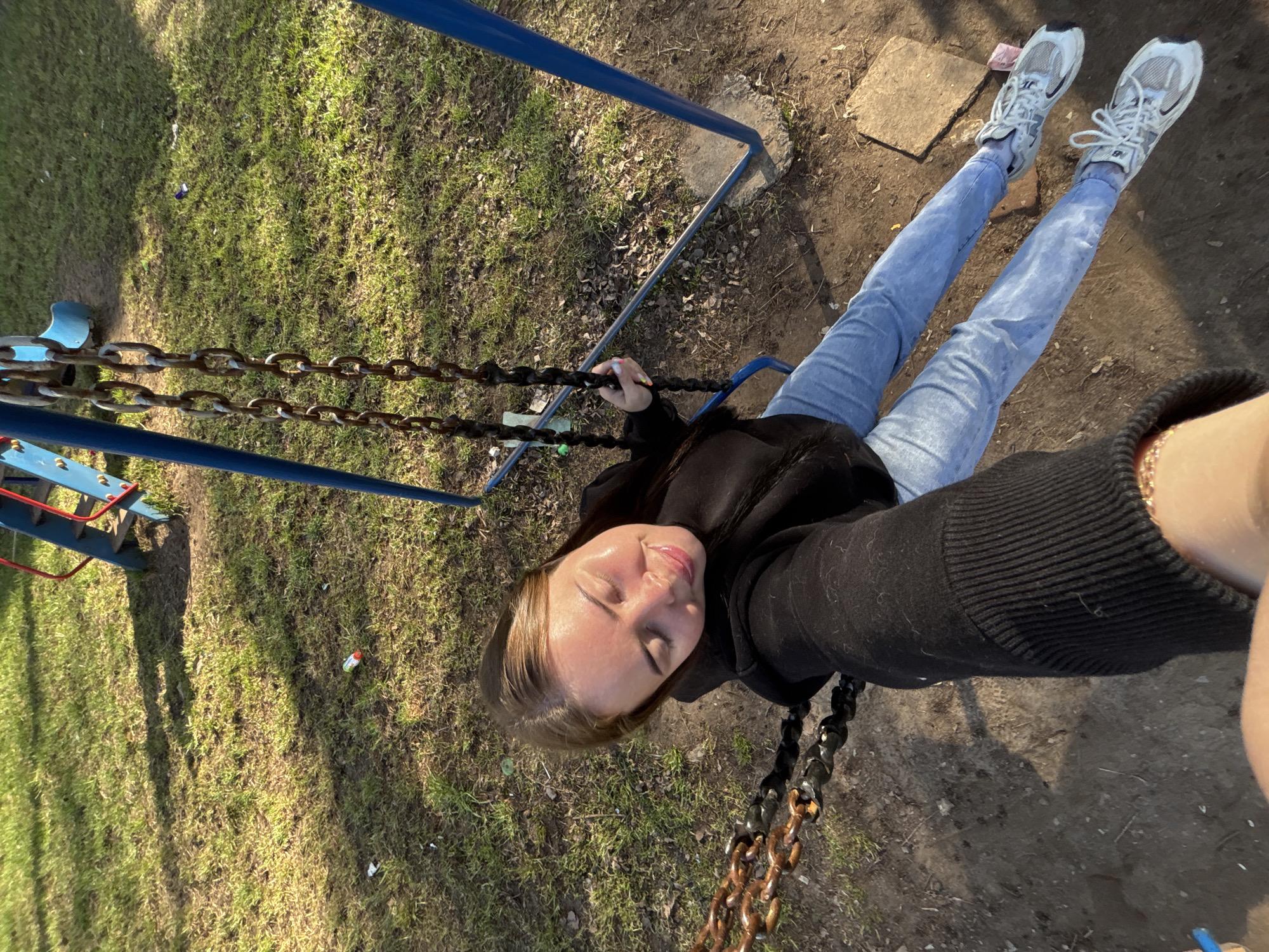 The image shows a woman sitting on a swing in a playground, likely in Izhevsk, Russia, based on contextual information not provided in the image. She is wearing light blue jeans, white sneakers, and a black hooded sweatshirt. She's holding the chains of the swing. The swing set has blue supports. The ground around the swing is covered in grass and some bare earth. The photo appears to have been taken from a high angle.
