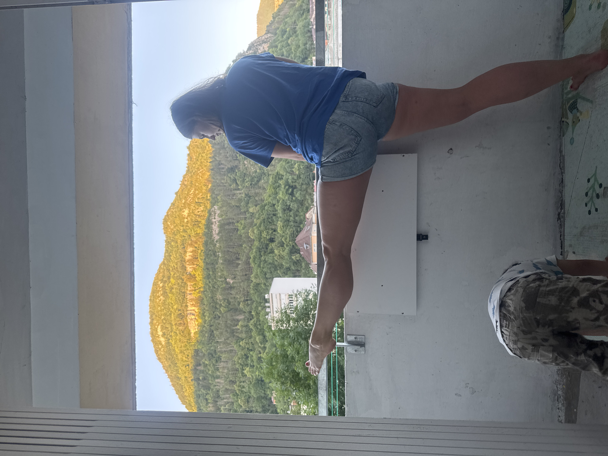 In Saint-Claude, France, on a balcony with a view of a mountain range and a town, a woman performs a split with her left leg extended horizontally. She is wearing a blue t-shirt and denim shorts. Below her, a child in camouflage pants and a white t-shirt is bent over, facing away from the camera. The balcony is concrete and has a railing.