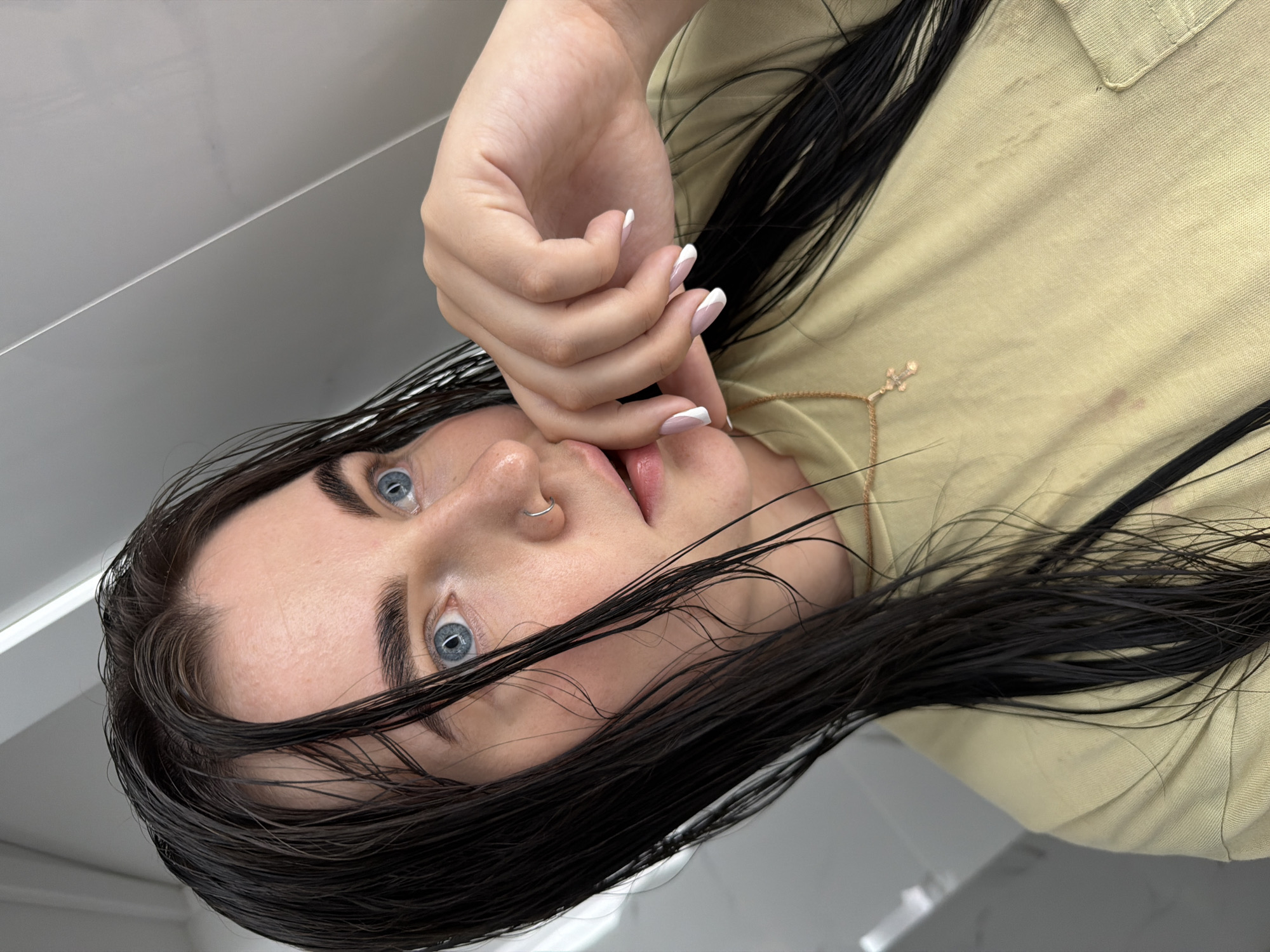 A young woman with pale skin and blue eyes is photographed in a close-up, upside-down perspective. Her long, black, wet hair is parted in the middle and falls around her face. She is wearing a light green shirt and a thin gold necklace. Her right hand is near her face, and she is wearing a French manicure. A silver nose ring is visible in her left nostril. The background appears to be a white, flat surface. The image does not provide enough information to confirm the location in Chaikovsky, Russia.