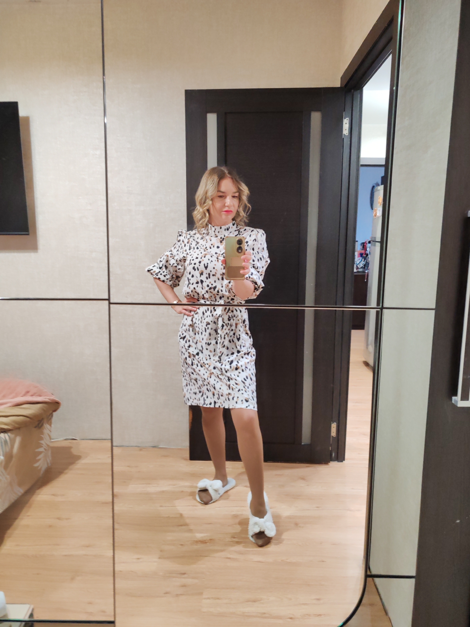 A woman is taking a selfie in front of a mirrored closet. She is wearing a white dress with black and brown spots and white slippers with bows. She has blonde, shoulder-length hair and red lipstick. She is holding a cell phone up to her face, and her right hand is on her hip. The closet has a horizontal bar across the middle of the mirror. Behind her is a dark brown door that is partially open. Through the doorway, a glimpse of another room can be seen. To the left of the closet is a bed with a pink and brown covering. The floor is light brown wood. This image was taken in Petropavlovsk-Kamchatsky, Russia.