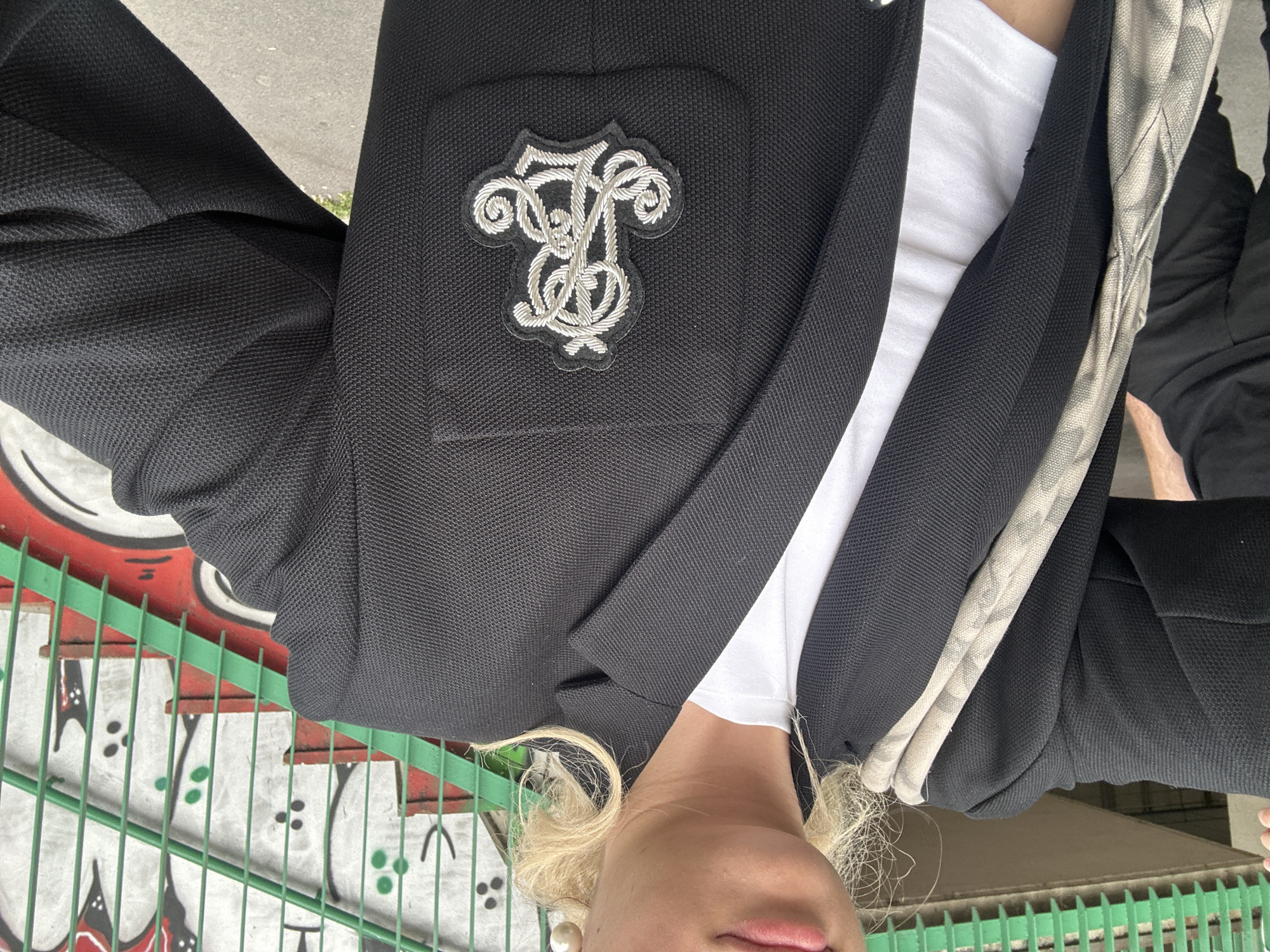 A medium-shot, angled photograph features a person in a black blazer with a white shirt underneath, posing in an upside-down orientation. The blazer has a detailed, silvery-white monogram patch on the upper left chest. The individual has blonde hair. The background includes a graffiti-covered wall with red, white, and black designs. A green metal railing runs across the lower portion of the image, partially obscuring the view. The context and style suggest the location is in the city of Milan, Italy, although this is only a contextual assessment based on the overall style of the photo.