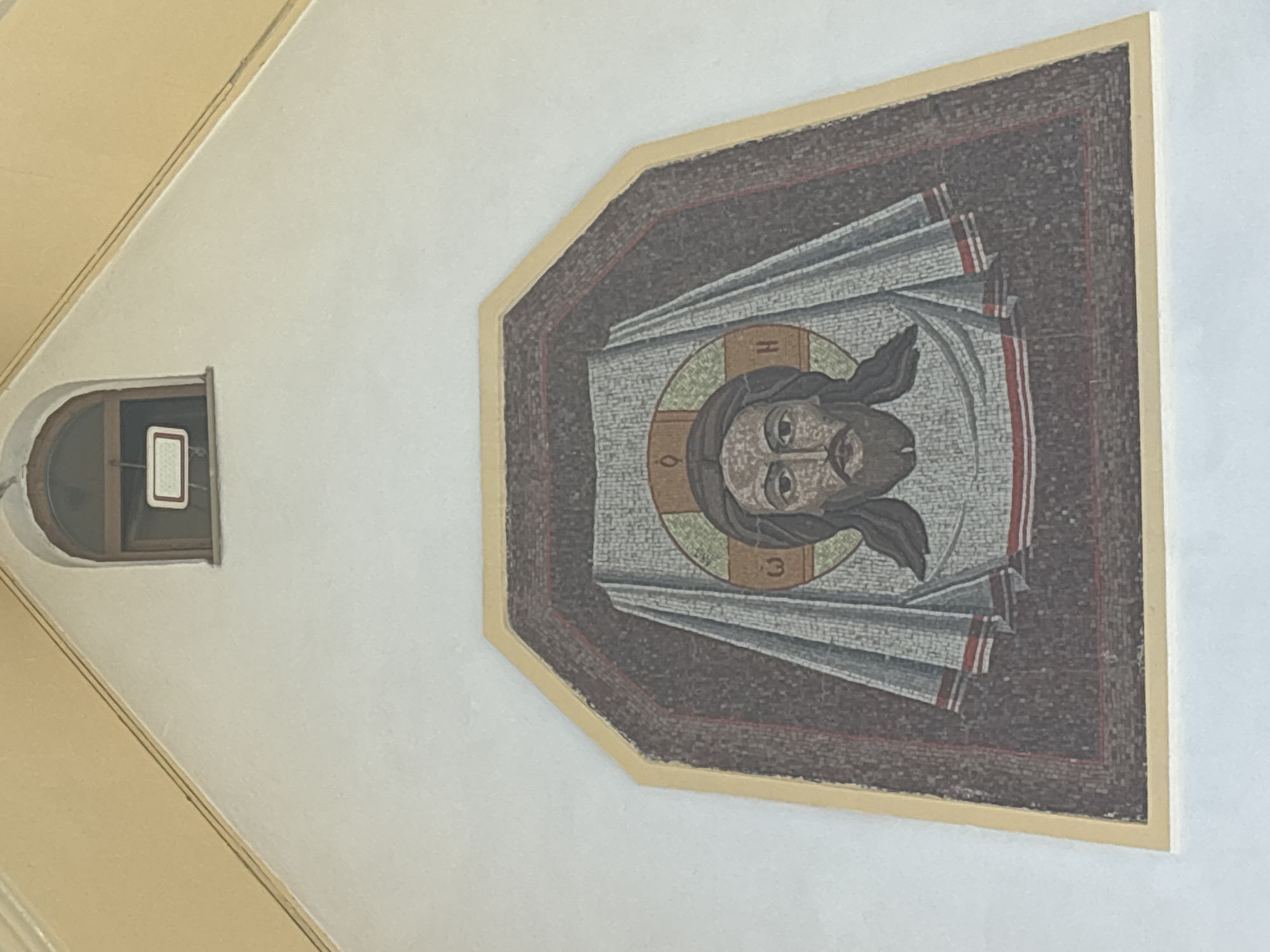 The image shows a mosaic depiction of Jesus Christ on the exterior wall of a building in Novosibirsk, Russia. The mosaic is framed by a light yellow border and depicts Jesus with a beard and long hair, wearing a draped garment. A halo surrounds his head, with Greek letters visible within it. Above the mosaic, a small, arched window with a wooden frame is present, containing a white rectangular sign with black text. The building's wall is painted white, and the roofline above the window is painted a light yellow. The overall setting appears to be an exterior facade, likely part of a religious or historical building.