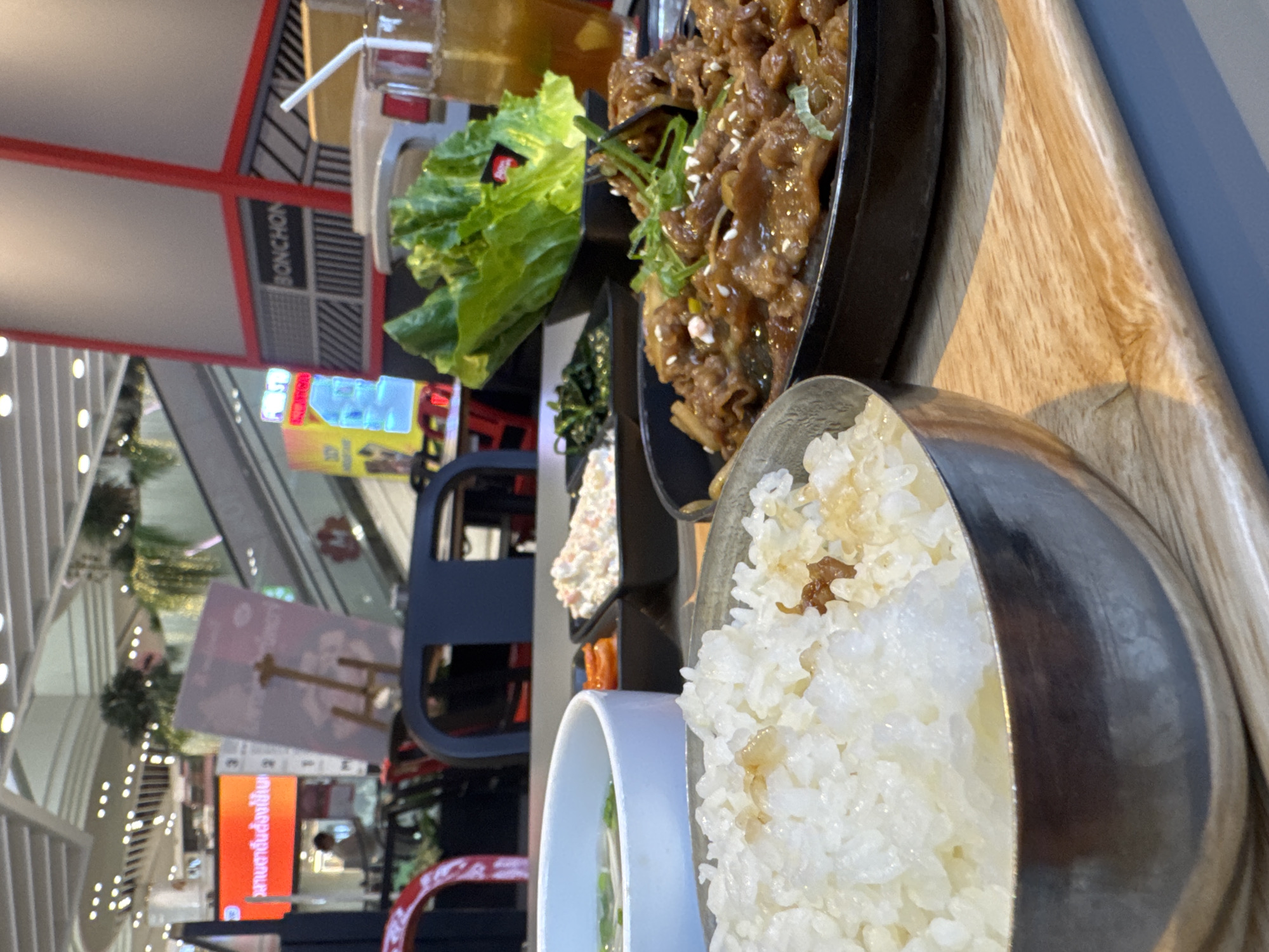 A close-up, high-angle shot showcases a meal at a Korean restaurant in Bangkok, Thailand. The foreground features a silver bowl filled with fluffy white rice, lightly seasoned. To the right, a dark, circular pan holds a serving of savory bulgogi, garnished with green onions and white sesame seeds. A bed of fresh green lettuce leaves sits in a black square dish beside the bulgogi, and next to that, a smaller black dish contains a white, creamy salad with colorful flecks of seasoning. A glass of iced tea with a straw and lemon slice completes the meal presentation. In the background, blurred elements of a mall environment are visible, including escalators, illuminated signs, and seating areas, suggesting the restaurant is located within a shopping center in Bangkok. The overall atmosphere is that of a casual dining experience.