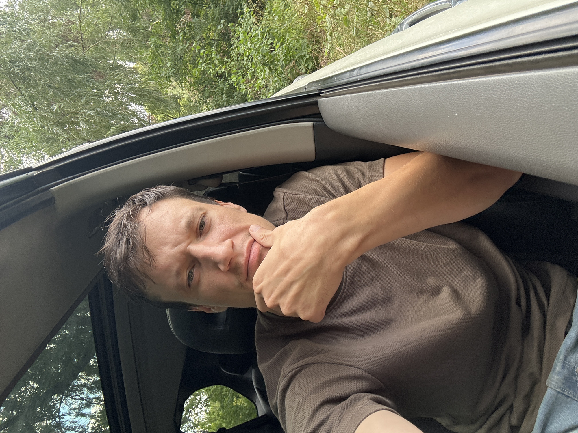 In the city of Makiv, Ukraine, a man is captured in a selfie taken from inside a vehicle, possibly a car. He is positioned in the driver's seat, with his right arm resting on the armrest and his hand supporting his chin. The man is wearing a brown, short-sleeved collared shirt and blue jeans. The interior of the vehicle is visible, including a sunroof through which sunlight filters. The background reveals a dense, green forest, suggesting the vehicle is parked or traveling through a rural or wooded area. The lighting is natural, indicating it's daytime, and the overall atmosphere appears relaxed and casual.