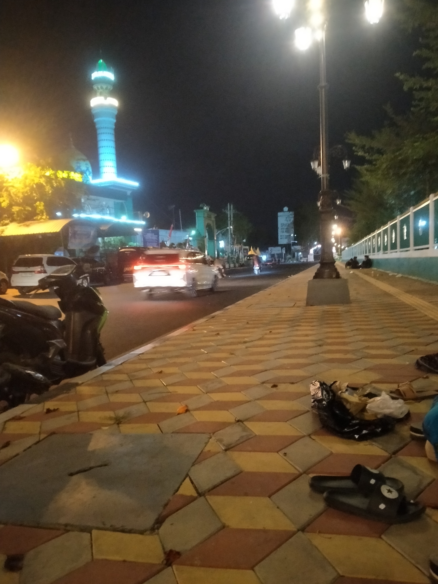This is a night time photograph taken in the city of Pamekasan, Indonesia, likely on Jalan Trunojoyo, a main street. The scene depicts a streetscape with a mosque on the left, characterized by its illuminated minaret and dome. A street light with multiple lamps stands prominently in the middle of the sidewalk. The sidewalk itself is paved with patterned concrete tiles in yellow and brown.
On the right side of the street, there is a white wall with green accents and vertical white bars. Several people are seated on the sidewalk near this wall. The street is active with vehicular traffic, including cars and motorcycles. One car in the foreground has its brake lights illuminated. There are also parked motorcycles and a white van on the left side of the street.
Scattered on the sidewalk are various objects, including a black plastic bag filled with debris, a pair of black sandals with white stars, and some fallen leaves. The overall environment is dimly lit by streetlights and the illuminated mosque, creating a nocturnal urban atmosphere.