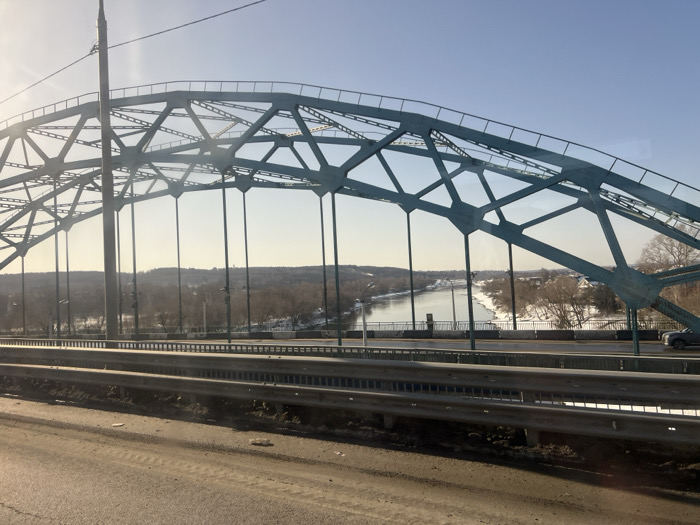 A wide exterior view captures a teal-colored steel arch bridge over a river in Zaozerye, Russia, under a clear, sunny sky. The bridge features an overhead truss structure with geometric patterns and vertical supports extending to the deck. A lamppost is visible on the left side of the bridge deck, partially silhouetted by bright sunlight from the upper left. The river below is bordered by snow-dusted banks and a distant treeline of mostly bare deciduous trees, indicating winter conditions. A dark-colored sedan or SUV is visible on the bridge deck, traveling from left to right. In the immediate foreground, a three-bar silver metal guardrail runs horizontally, separating the viewer's elevated position from the bridge deck. The paved surface in the foreground is soiled with dirt and debris. No individuals are present in the scene.