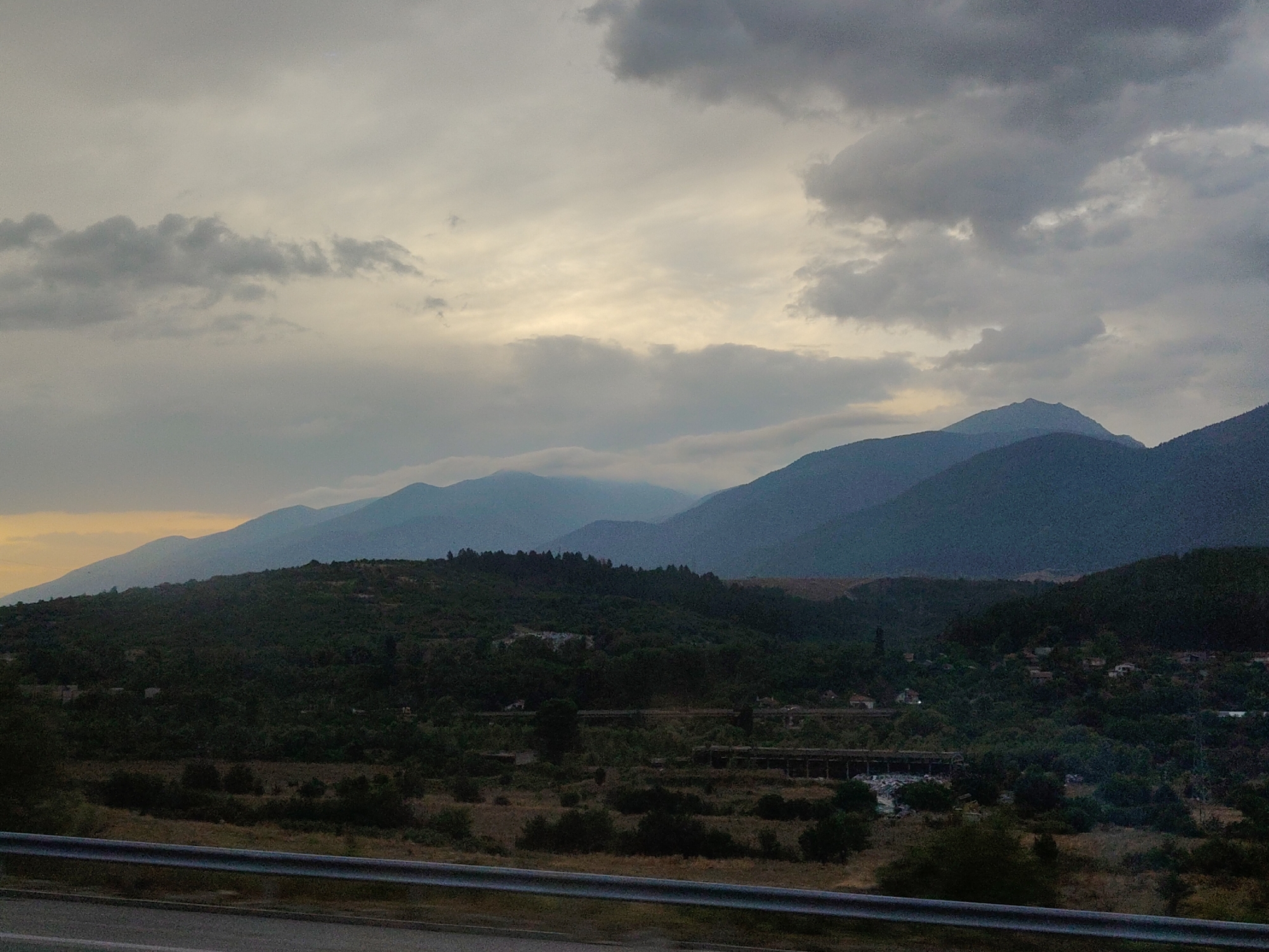 The image depicts a rural landscape with layered mountains and dense vegetation under a cloudy sky. In the foreground, a metallic barrier and a road suggest a viewpoint from a highway. The middle ground is characterized by rolling hills covered in dark green trees, with scattered buildings and evidence of some infrastructure, possibly remnants of construction or a bridge, visible amidst the foliage. The background features a series of blue-hued mountains, fading into the overcast sky. The overall atmosphere is somber, hinting at twilight or overcast weather. The scene is not specific to the city of Djerman, Bulgaria, as no identifying landmarks or characteristics are present to confirm its location within that city.