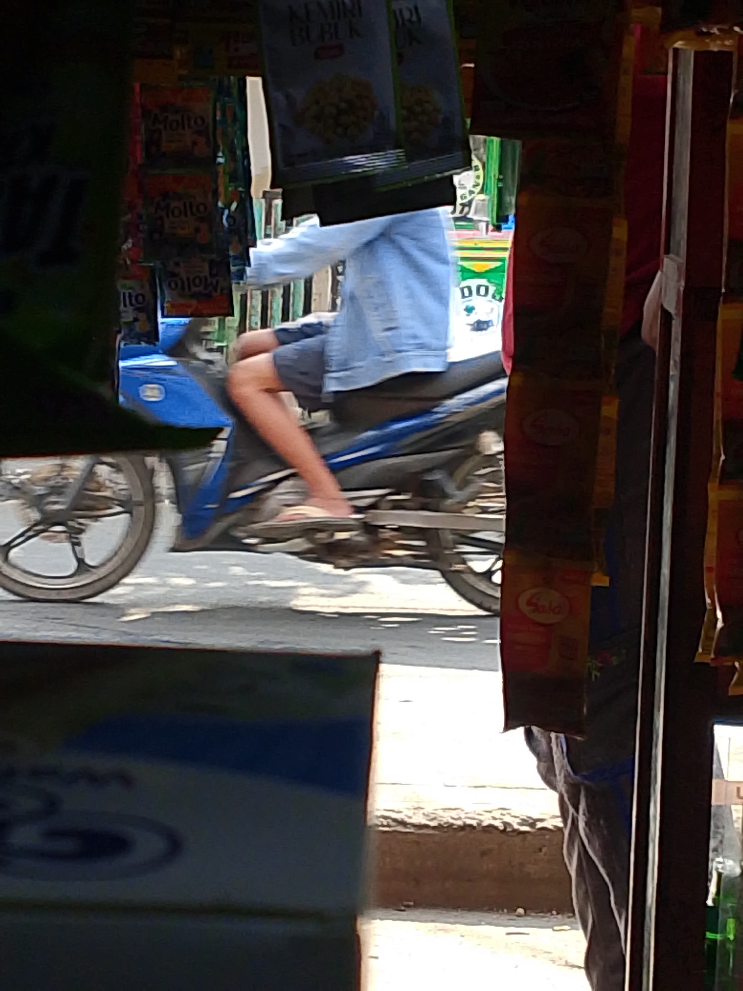 The image captures a street scene in Palembang, Indonesia, with a person riding a blue motorcycle. The motorcycle's front wheel and part of the frame are visible on the left, while the rider, wearing a light blue denim jacket and grey shorts, is in motion. The rider's legs are extended, and they are wearing sandals. The motorcycle appears to be traveling on a paved surface, possibly a road or sidewalk.
In the background, a small stall or shop is visible, with various products displayed. Several packets of what appear to be food or detergent are hanging from the stall. Some of these packets have visible branding, including "Molto" and "KEMIRI BUBUK". The stall has a rustic appearance, with wooden structures. The overall lighting suggests it is daytime.
To the right of the motorcycle and rider, there is a dark vertical element, possibly a doorway or a structural support of the stall. Further to the right, more packets and products are displayed, including a green bottle which might be a beverage. In the foreground, a portion of another object, possibly a box or a shelf, is visible, out of focus and partially obscuring the view. The environment seems to be an outdoor marketplace or a small roadside shop.