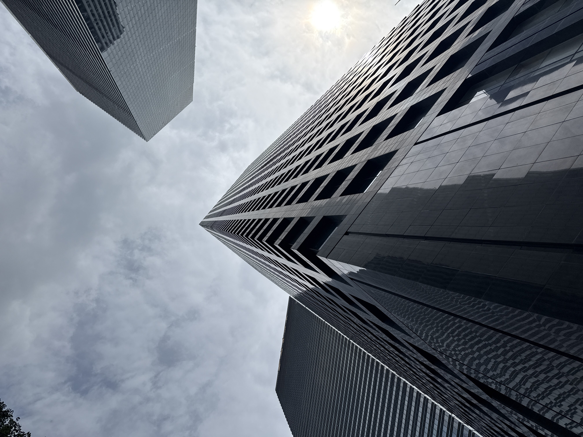 The image is a low-angle shot of modern skyscrapers in Tianhe District, China, against a cloudy sky with a visible sun flare. The foreground features a dark, reflective skyscraper with a grid-like pattern of windows and dark paneling. To the left, another skyscraper with a ribbed facade is visible. In the background, a lighter-colored skyscraper with a similar grid pattern is also present. The overall scene depicts an urban environment with towering architectural structures. There are no people or identifiable actions visible.