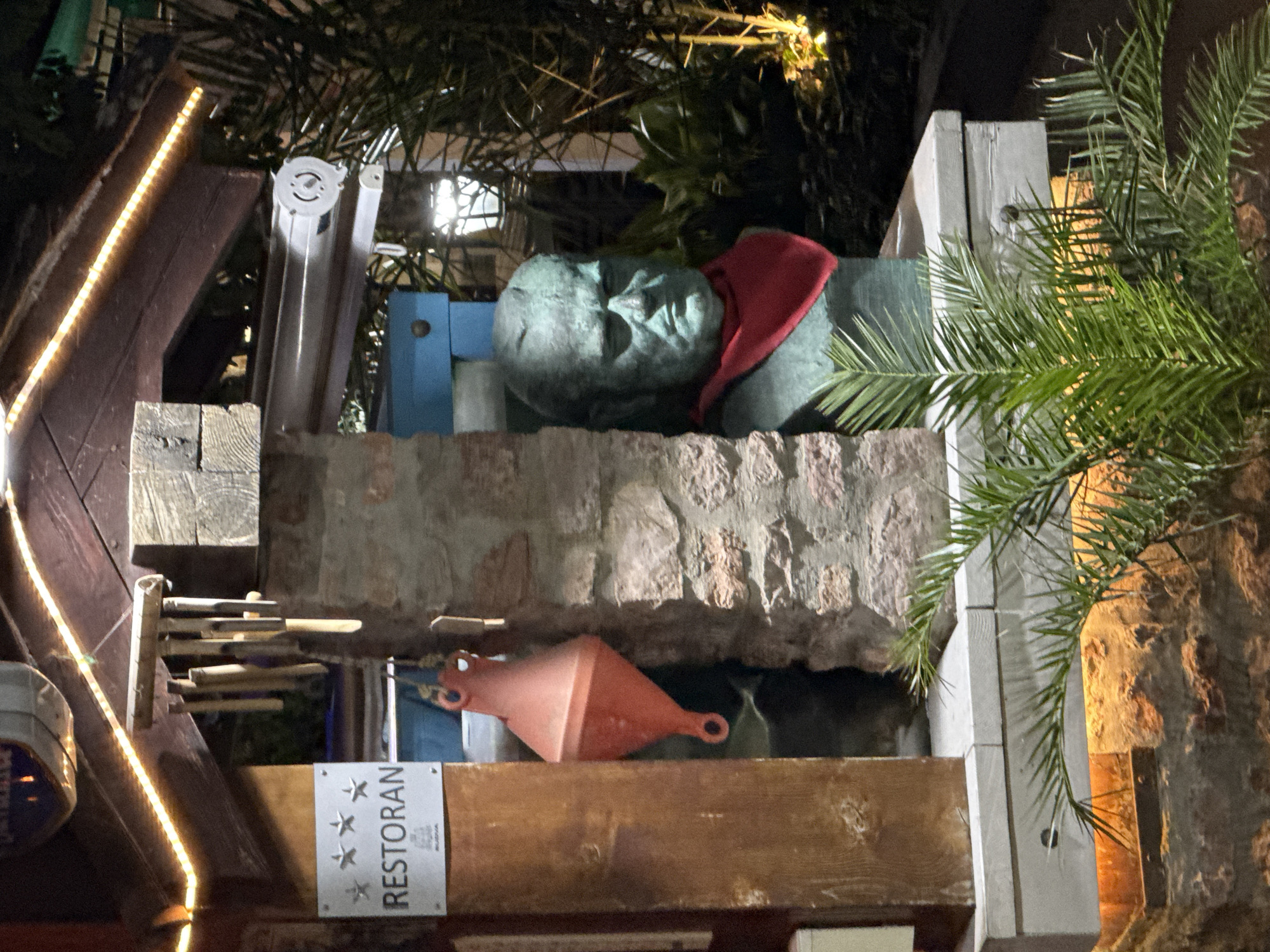 The image shows a bust of a man wearing a red scarf, displayed on a stone wall in Budva, Montenegro. To the left of the bust, a sign reads "RESTORAN" with three stars above it. A red buoys hangs from a string next to the sign. The scene appears to be outdoors, possibly at night, with string lights illuminating the area. Plants and palm trees are visible in the background.