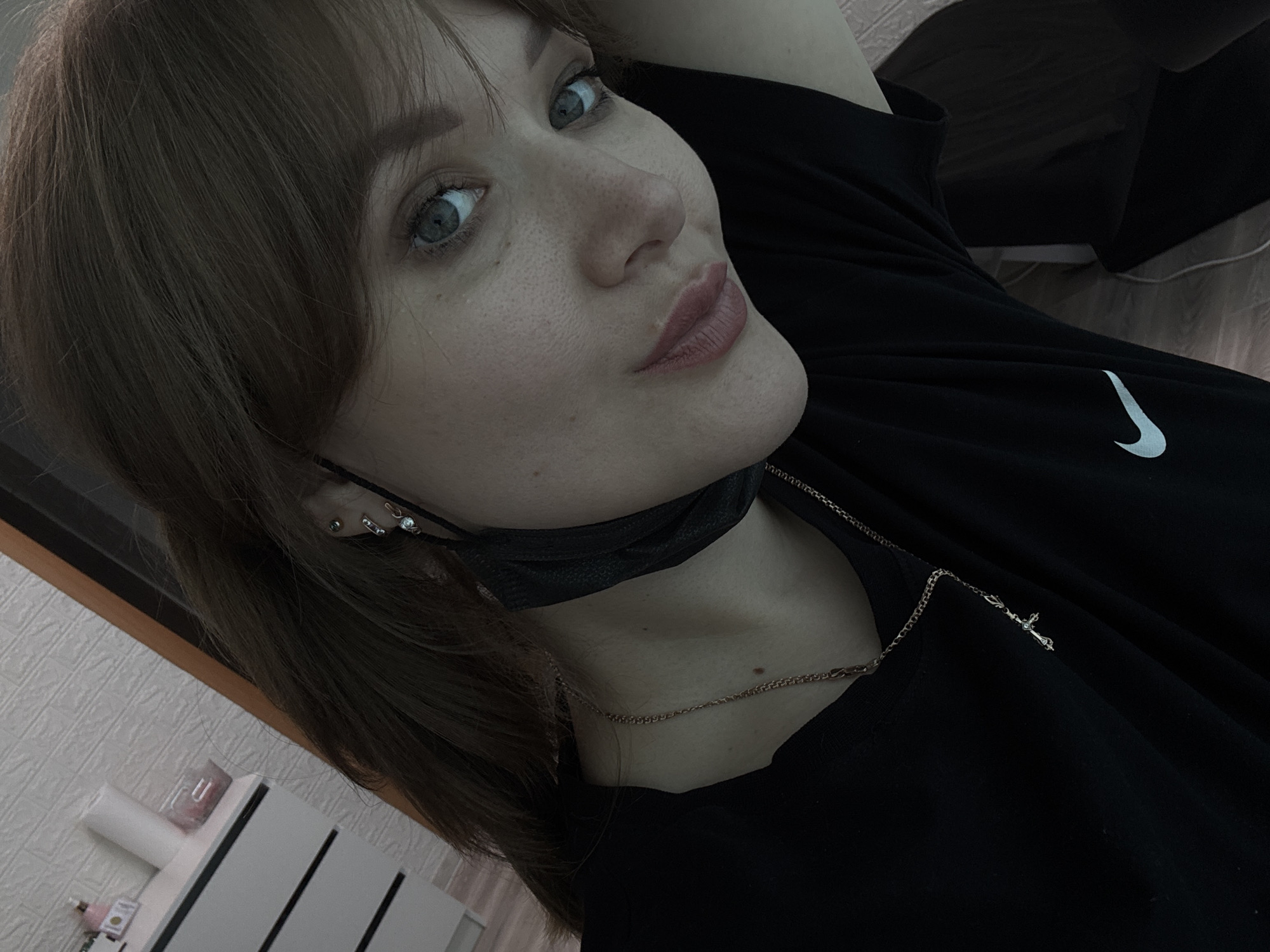A young woman with auburn hair and blue eyes is taking a selfie in a room. She is wearing a black t-shirt with a white Nike logo and a black face mask around her neck. A delicate necklace with a cross pendant is visible. The background suggests an indoor environment with a dresser or cabinet to the left and a dark curtain or doorway to the right. There are no indications of the city of Chaikovsky, Russia.