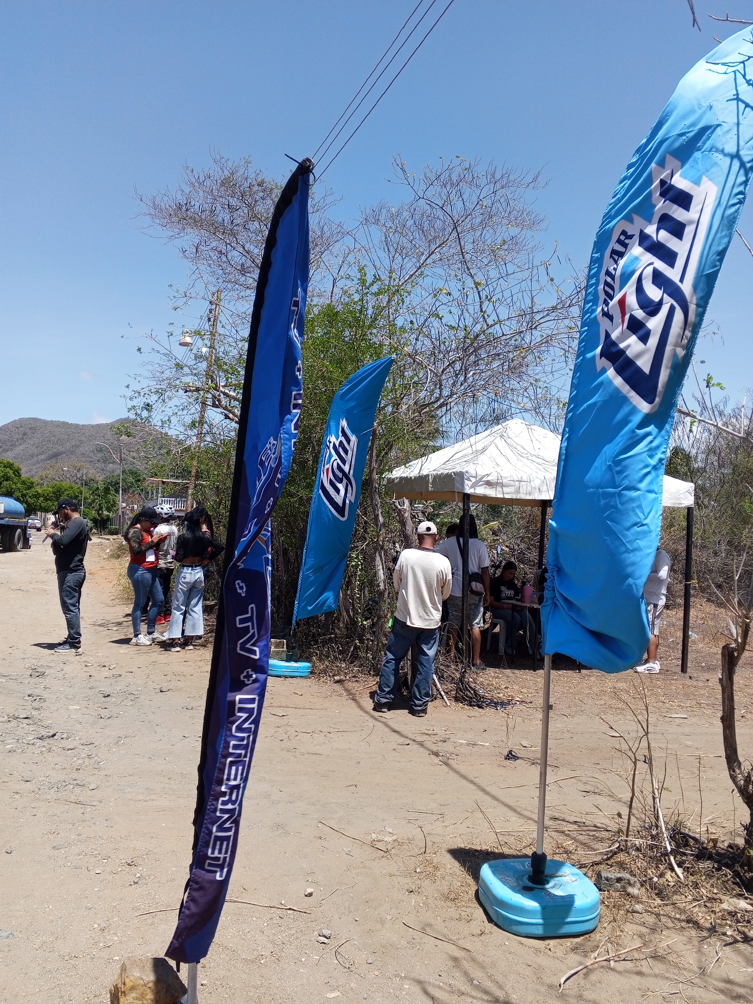 The image depicts a roadside scene in an area identified as Unknown, Unknown, with several notable elements. In the foreground, three tall, blue advertising flags stand out. One displays the "Polar Light" logo, another is partially obscured but shows "TV + Internet", and the third has a less distinct design. These flags are anchored with light blue plastic bases and rocks to hold them down.
In the background, a group of people gathers under a white tent-like structure. The individuals vary in attire, with some standing and others seated. To the left of the tent, a small group of people stand near the road, some appearing to be engaged with their phones. A man in a dark shirt stands slightly apart from the group, also looking at his phone. In the distance, a mountain range is visible under a clear blue sky.
The environment is dusty, with dirt roads and sparse vegetation, hinting at an arid climate. Power lines are visible overhead, and a truck parked to the left adds to the roadside setting. The overall impression is a casual, outdoor gathering or event occurring on a sunny day in a somewhat remote location.