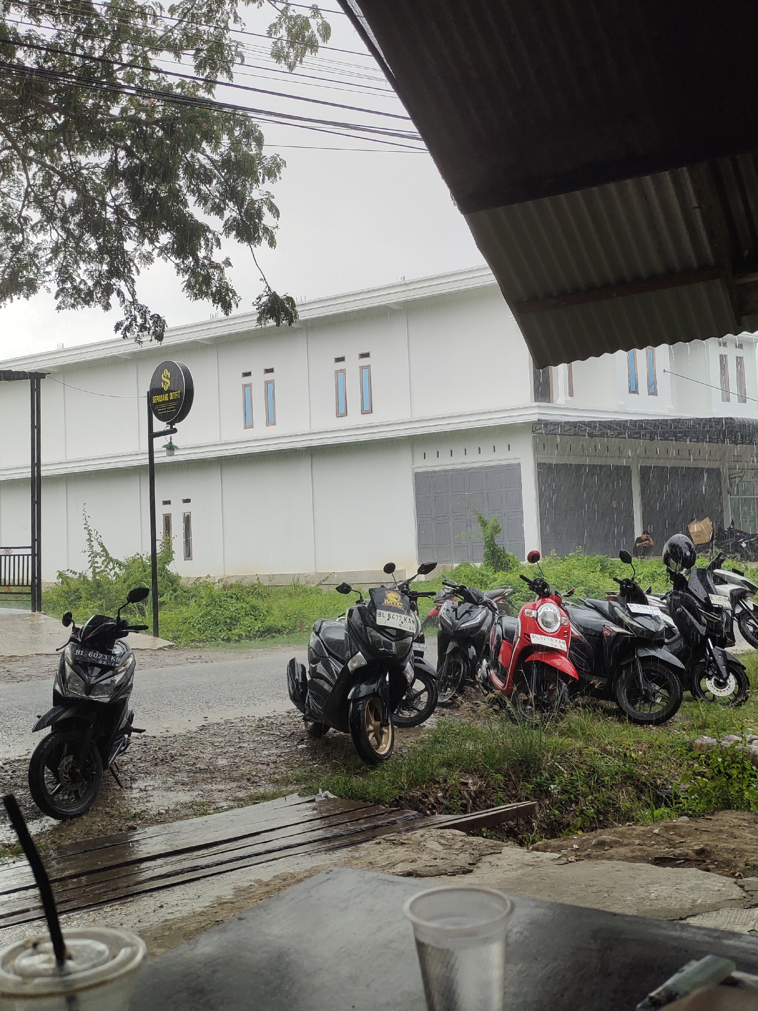 The image depicts an outdoor scene on a rainy day in Keudematangkuli, Indonesia. In the foreground, a wooden platform is visible, with two plastic cups and a straw on it, suggesting a beverage has been or is being consumed. Beyond the platform, a wet asphalt road is lined with several parked motorcycles. The motorcycles are of various colors, including black and red, and some have license plates.
Behind the motorcycles, a patch of green grass and foliage grows, with a small ditch or water channel running through it. Further back, a large, white, two-story building stands, with several windows and a large garage-like entrance on the ground floor. A black sign with a circular logo containing a stylized "S" and the text "SEPASANG OUTFIT" is mounted on a pole in front of the building. The sky is overcast, and rain is visibly falling, creating streaks in the image. Trees with green leaves are present in the upper left portion of the frame, and power lines are visible crisscrossing the sky. The overall impression is one of a quiet, somewhat dreary day, with the rain as a prominent environmental factor.