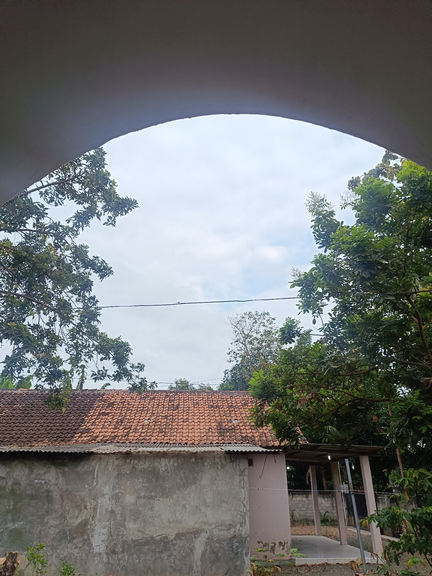 The image shows a rural scene in Bantul, Indonesia, looking up through a curved archway towards a cloudy sky. The archway is a light gray, possibly concrete or plaster. Below the sky, to the left and right, are dense green trees with dark green leaves. The trees frame a view of a single-story building with a red tile roof and a corrugated metal awning. The building's walls are a rough, light gray stucco on the left, and a pale pinkish hue on the right, with visible support columns. A thin black wire stretches horizontally across the sky. The overall impression is of a residential area or a small compound in a tropical climate.