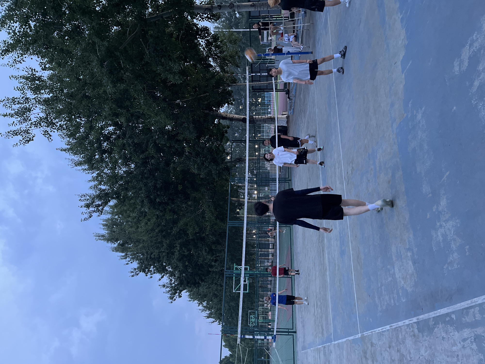 This media file captures an outdoor scene of a group of people playing volleyball on a court. The court is blue with white lines marking boundaries, and a white net divides the playing area. A volleyball is visible in the air, suggesting an active game is in progress.
Several individuals, dressed in casual sportswear, are positioned on the court, with some in motion. One person in the foreground wears a black long-sleeved shirt and black shorts, with white socks and athletic shoes. Another person, further back on the same side of the net, is in a white t-shirt and black shorts. On the opposite side of the net, people are also dressed in athletic attire, including a person in a light grey t-shirt and black shorts, and another in a white t-shirt and black shorts. In the background, more people can be seen watching or participating, and there is a basketball hoop visible behind the volleyball net.
Lush green trees border one side of the court, providing a natural backdrop. The sky is visible, appearing overcast or twilight, suggesting it might be early morning or late evening. The lighting is subdued, consistent with these times of day. The overall setting appears to be a park or recreational area. There is no visible text in the image. The activity is clearly a friendly game of volleyball.