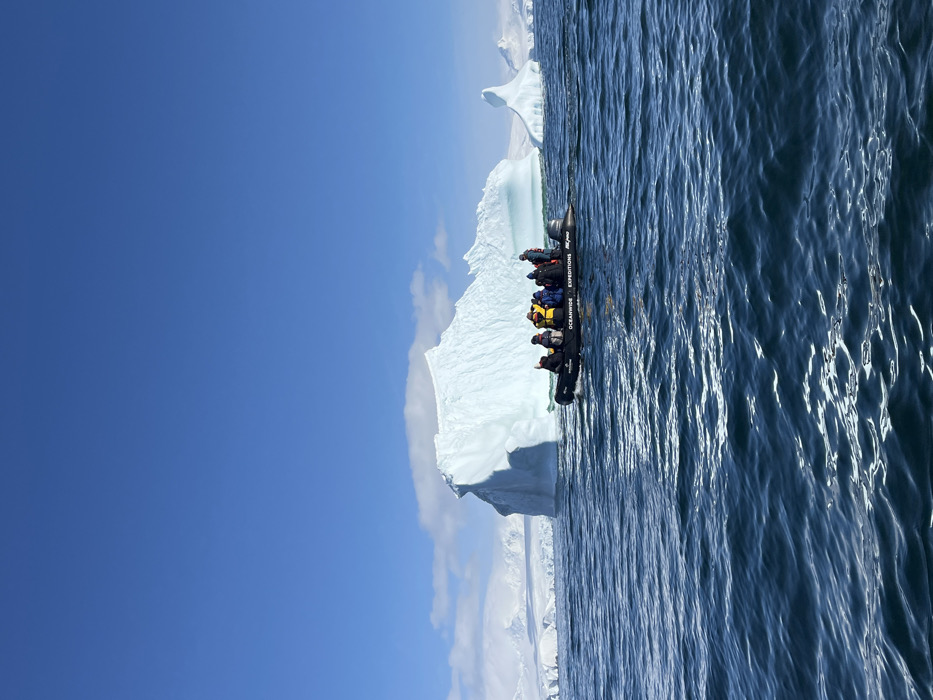 The scene depicts a polar maritime environment, not an urban location. A black rigid-hull inflatable boat (RIB), marked with "OCEANWIDE EXPEDITIONS" and partially visible "AK-PRO" in white text on its side, moves across dark blue, rippled water from left to right. Approximately ten individuals are seated in the boat, clad in heavy winter expedition gear including multi-colored parkas (yellow, red, blue) and black life vests. One passenger on the port bow has their right arm raised. In the mid-ground, a substantial, irregularly shaped iceberg of glacial ice exhibits white and blue hues with distinct shadowed facets. To the right of the primary iceberg, a smaller, conical iceberg is visible. The distant background features snow-covered mountain ranges and glaciers beneath a clear, bright blue sky with minimal scattered white clouds along the horizon.
