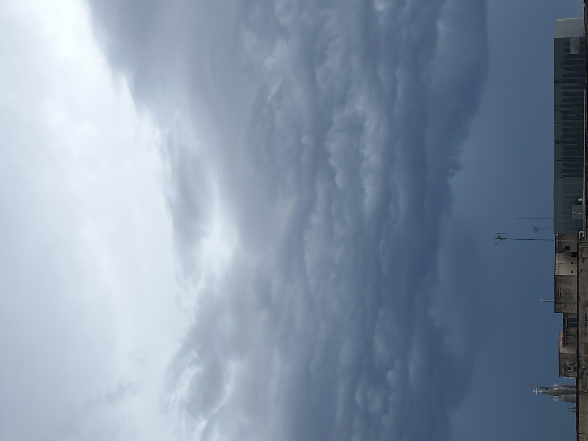 The image depicts a view of the sky in Barcelona, Spain, with prominent cloud formations. The sky is predominantly gray, suggesting overcast conditions. A tall building is visible on the right side of the image, with antennas on the roof. The building's architecture is modern, indicating an urban setting. There are no visible people, actions, or interactions within the frame. The environment suggests a cloudy day in an urban area.