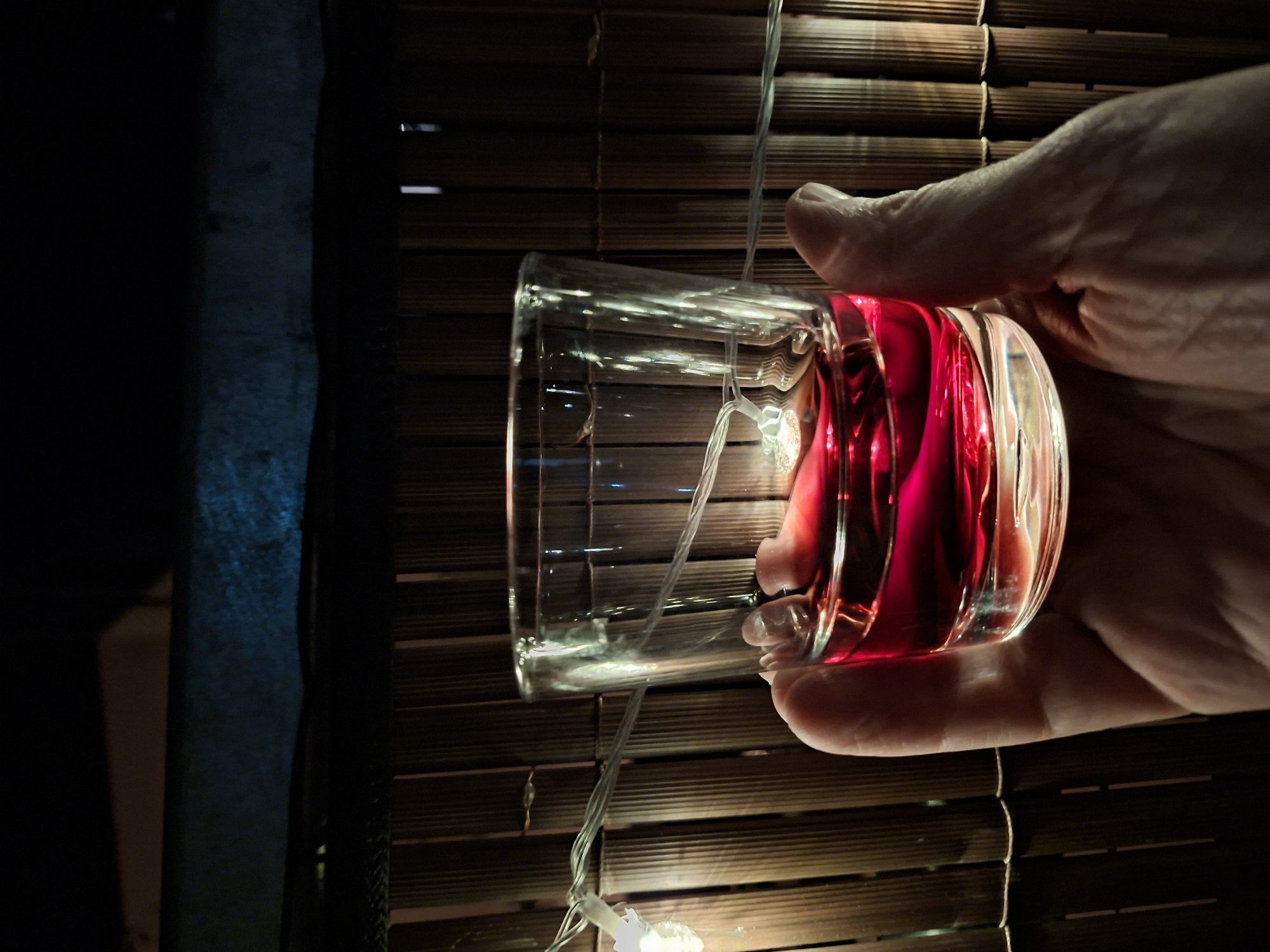 A hand holds a clear glass containing a red liquid, possibly wine or a cocktail. The glass is illuminated by string lights wrapped around it and behind it, creating a warm glow. The background consists of a woven bamboo fence, typical of outdoor patios or garden decorations. Above the fence, a dark blue illuminated surface is visible. The scene appears to be set outdoors at night. The specific location in Sinzheim, Germany, is not discernable from the visual information provided.