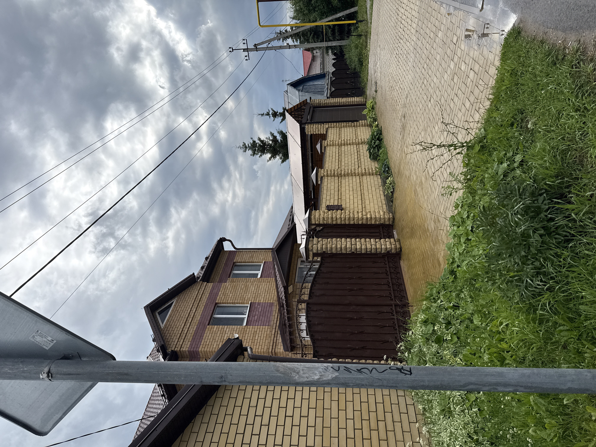 This is a wide shot, eye-level, outdoor, daytime photograph of a street in Kazan, Russia. The foreground features lush green grass on the left and a paved driveway on the right, with puddles indicating recent rain. A grey metal pole, likely a street sign, stands prominently in the left foreground, with graffiti scrawled on it. Behind the pole is a light brown brick house with a dark brown roof and a matching decorative metal gate. The gate leads to a driveway area that also appears to be paved. Further down the street, a brick wall extends, topped with a covered carport or garage structure with a transparent roof. Beyond that, several other houses and structures are visible, including a power line pole with electrical wires strung overhead. The sky is overcast with dark, dramatic clouds. The overall impression is of a residential street on a cloudy, possibly post-rainy day in an urban or suburban setting.
