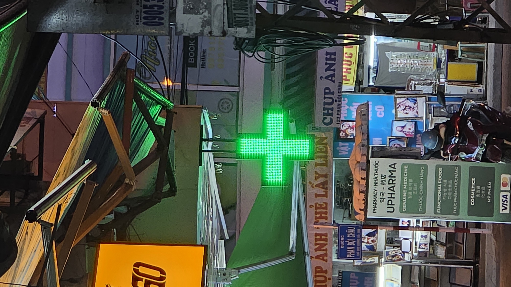 The image depicts a street scene in Nha Trang, Vietnam, featuring a pharmacy identified by a bright green, illuminated cross. The pharmacy signage displays the name "UPHARMA" along with various details in Vietnamese, Korean, and English, including the categories of products offered: "Genuine Medicine," "Functional Foods," and "Cosmetics." Additional signage includes the text "CHUP ANH THE LAY LIEN" and "CHUP ANH," indicating photo services.
The background consists of building facades and various signs and advertisements. A parked motorbike is partially visible on the right. In the foreground, there are scaffolding-like structures and additional signage, partially obscuring the view. Overhead, a network of wires is visible. The image has a nighttime ambiance.