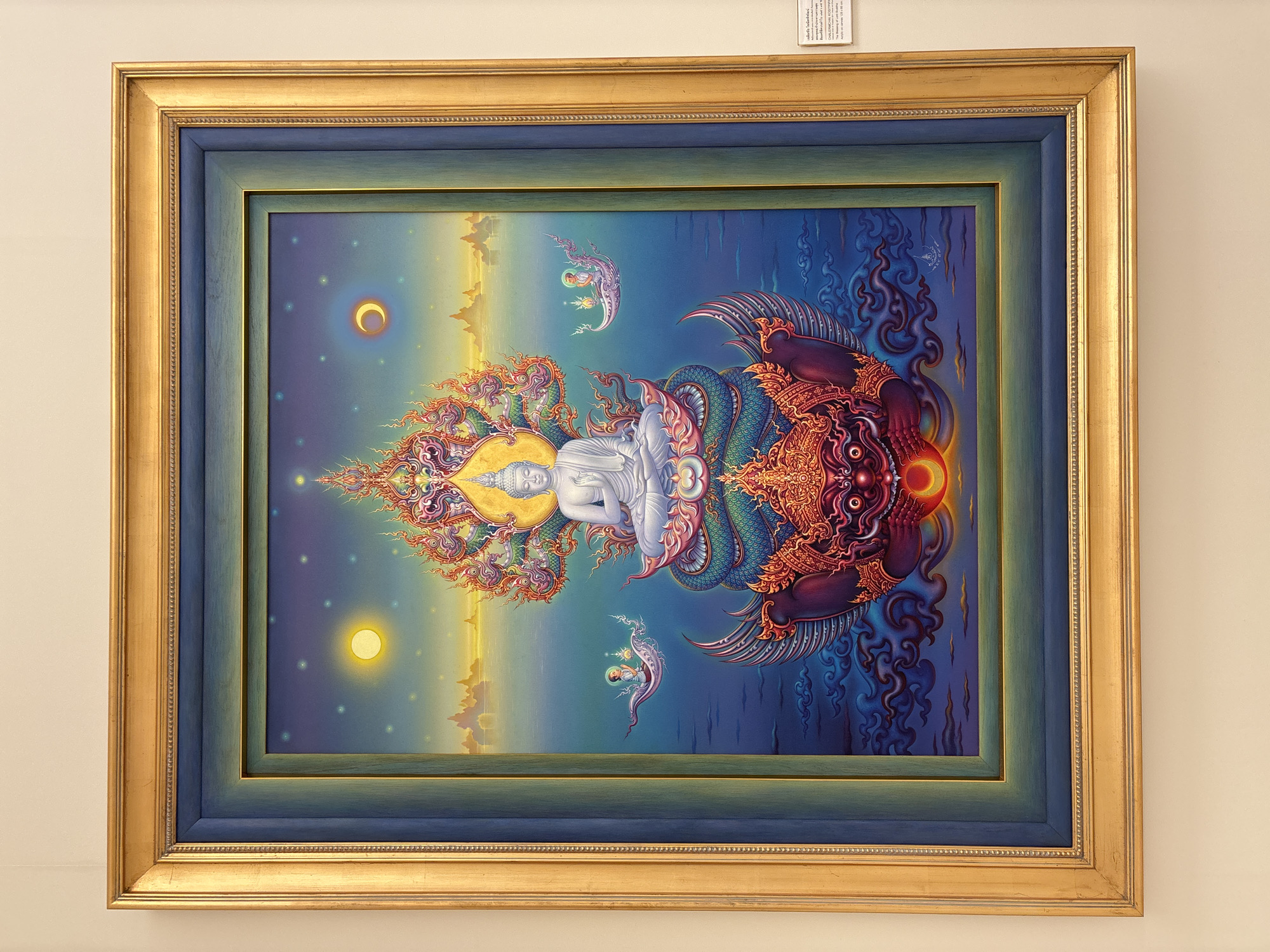 A framed painting hangs against a plain, light-colored wall. The painting depicts a reclining white Buddha figure centered between a golden crescent moon on the left and a bright sun on the lower left. Intricate colorful designs and ethereal beings surround the Buddha. To the right of the Buddha is a complex, multi-colored creature with large wings and a menacing expression. Blue and dark blue tones dominate the right side of the painting, contrasting with the yellow and golden hues on the left. The painting has a double frame, an inner dark blue frame and an outer ornate gold frame. In the upper right corner above the painting, a partially visible white information sheet is attached to the wall. The painting is likely located in a temple or art gallery. The subject of the artwork suggests a connection to Thai Buddhist art. While it is plausible that it is located in Bangkok, without further context, the precise location cannot be determined.