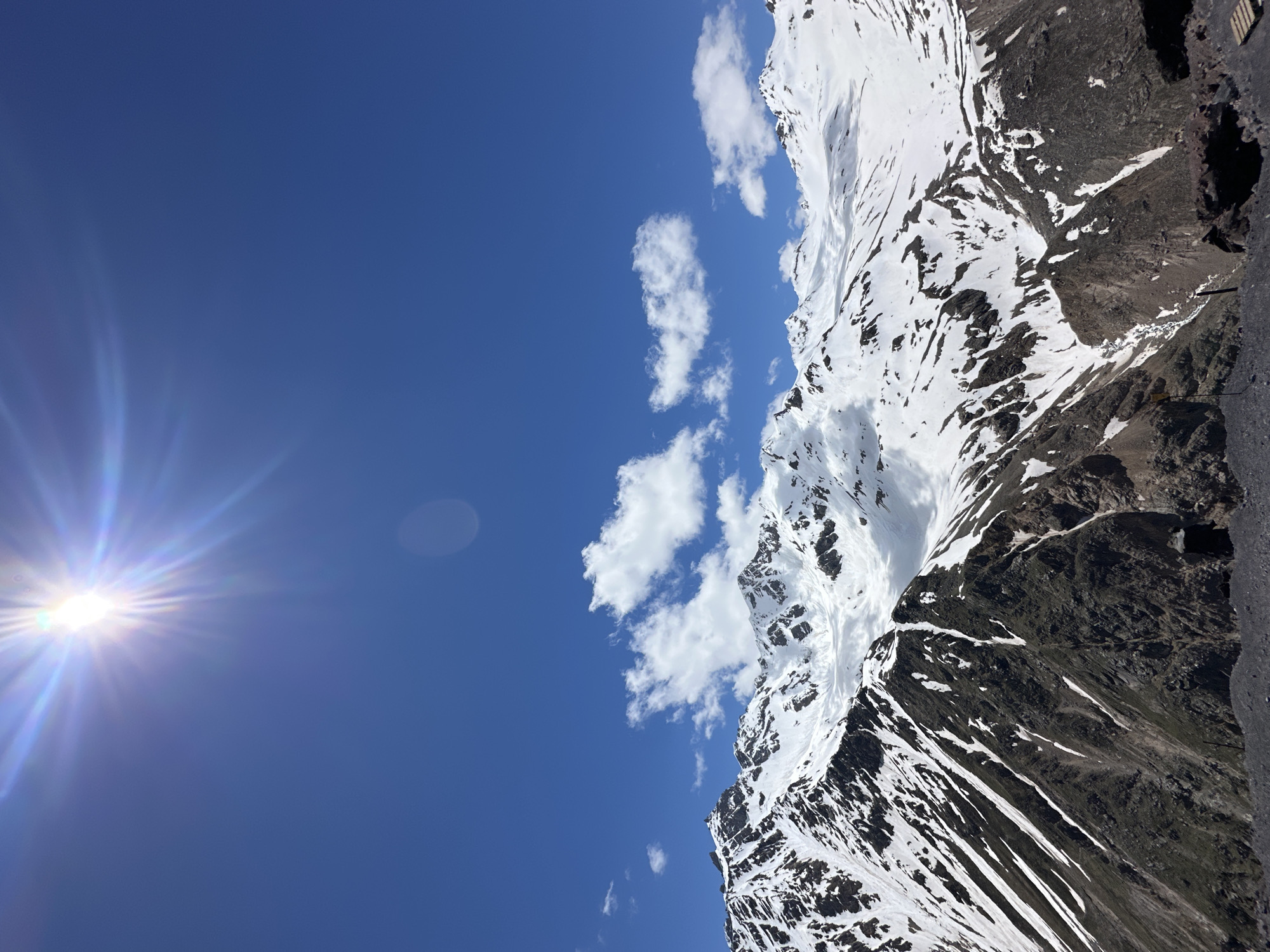A bright sun with a lens flare dominates the upper left portion of the image against a clear blue sky. Below, a mountain range with snow-capped peaks and rocky, darker slopes stretches across the frame. Patches of snow are visible on the slopes, with some areas appearing to be glaciers. Wispy white clouds are scattered across the sky above the mountains. The foreground consists of dark, rocky terrain. There are no visible people or active interactions in the scene. The image captures a mountainous, possibly alpine, environment under a bright sunny sky. The location is identified as Terskol, Russia.