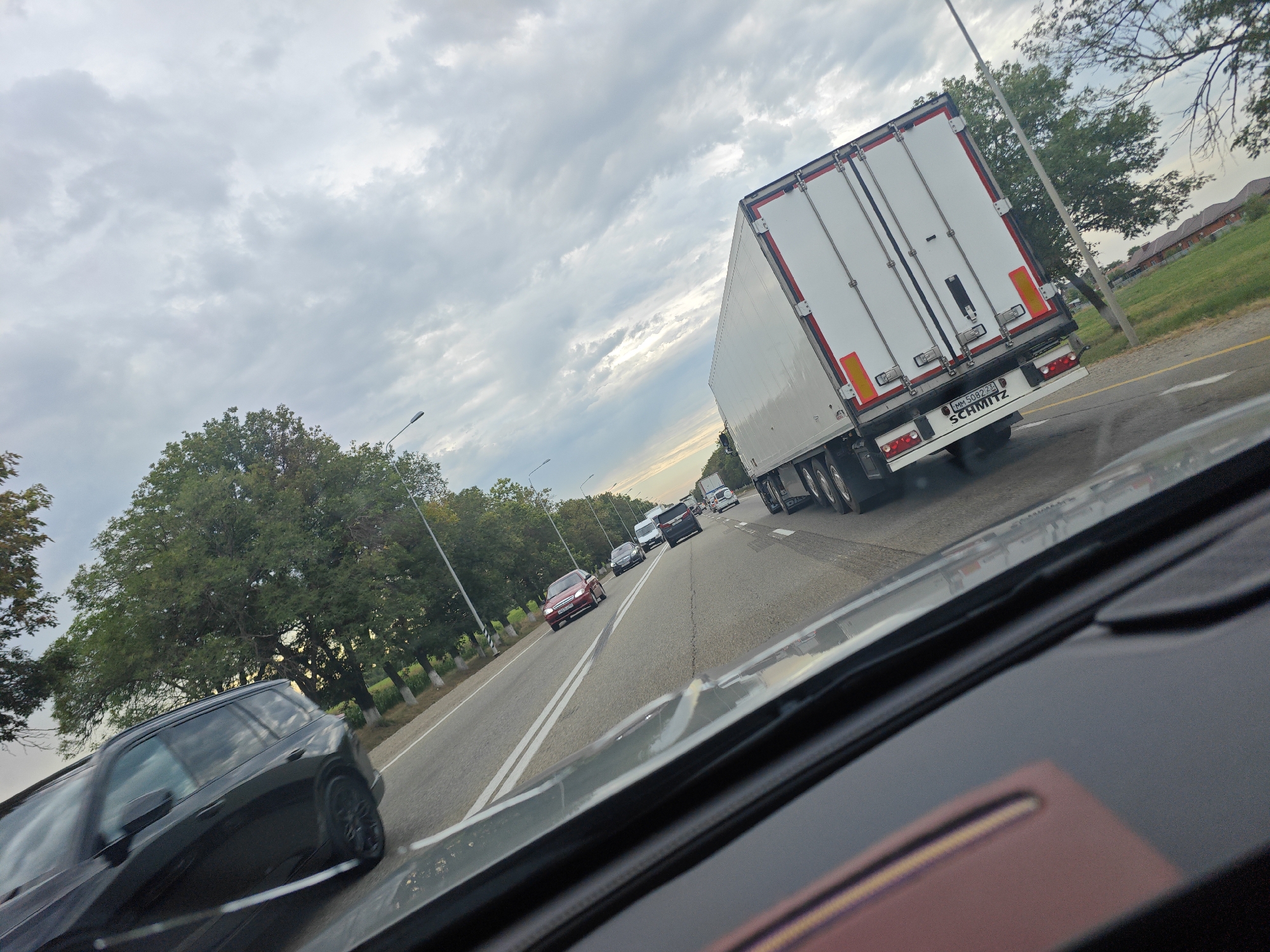 A wide-angle, eye-level shot captures a highway scene with multiple vehicles on a cloudy day. In the foreground, the interior of a car, including the dashboard and windshield, is visible. The main focus is on a large white semi-trailer truck with a red and white striped trailer, labeled "SCHMITZ," driving in the right lane. Several other cars are also on the road, including a black SUV in the left lane and a red car further ahead. Trees line the side of the highway, and houses are visible in the background. The road is paved and marked with white lines. The weather appears overcast. The location is identified as Zavetny, Russia.
