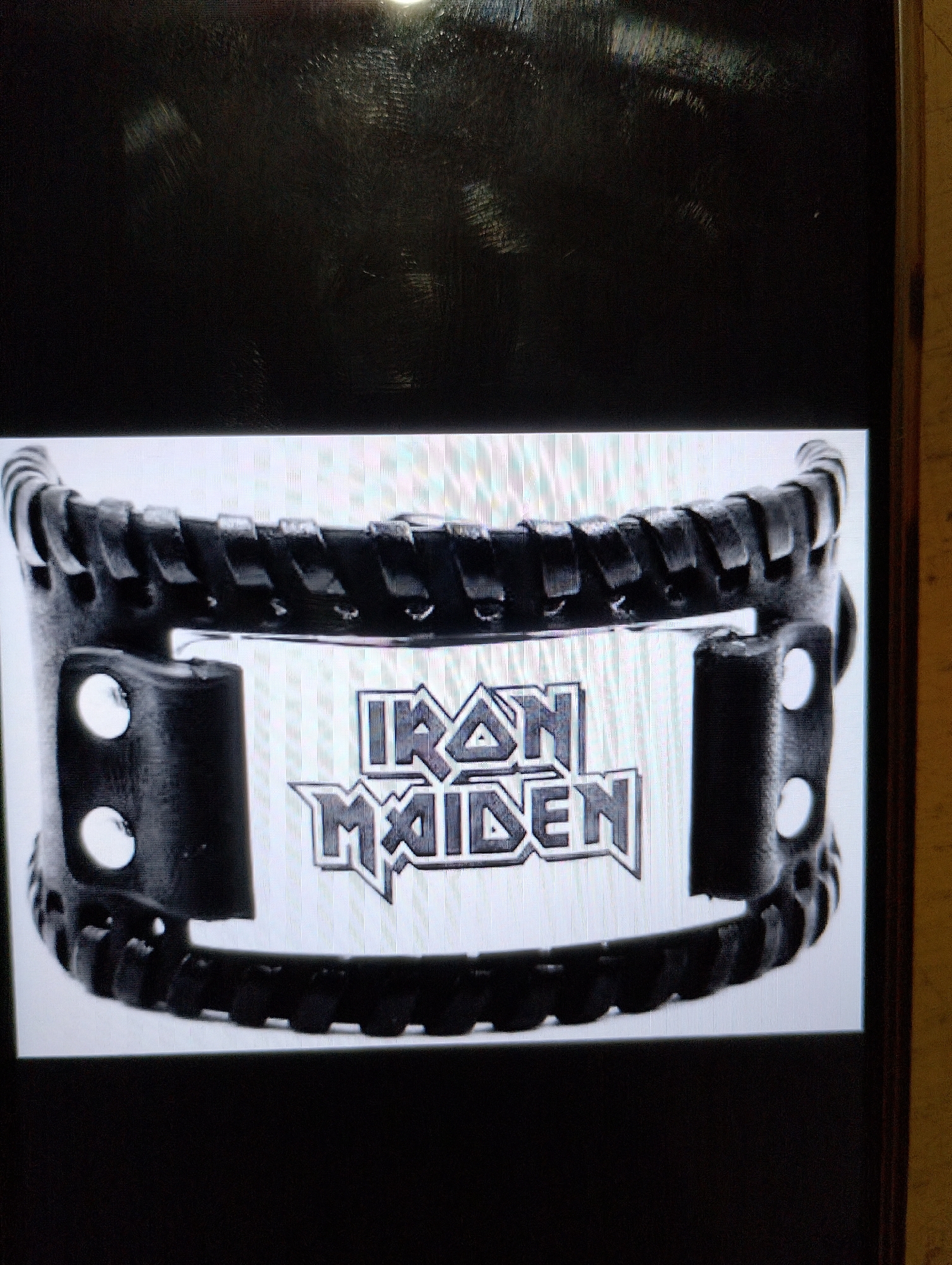 A black leather wristband with a metal plate is shown. The metal plate has the words "IRON MAIDEN" embossed on it in a gothic font. The wristband appears to be secured with two rivets on each side. The background is dark and out of focus.