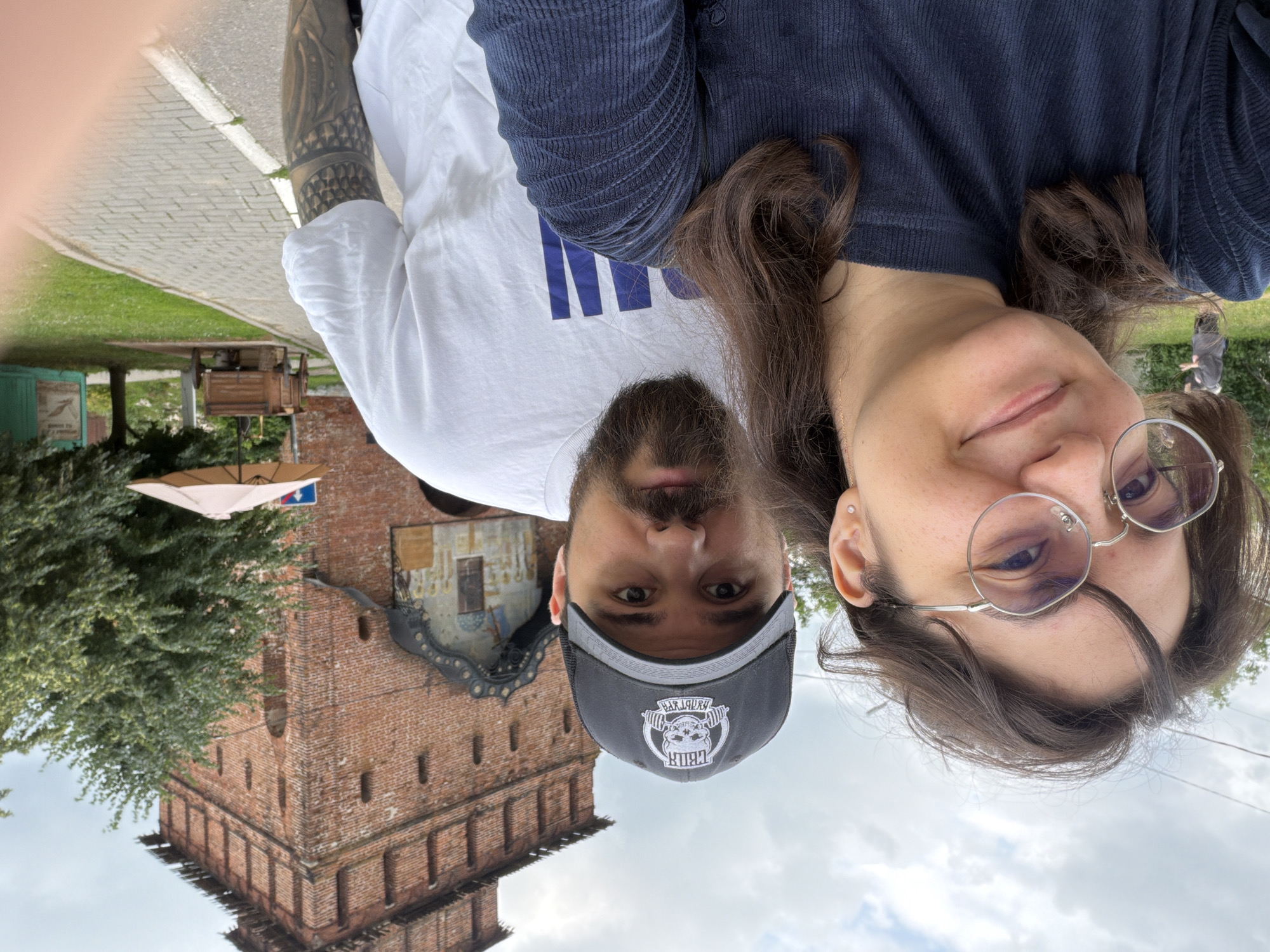 In Kolomna, Russia, two individuals, a woman and a man, are captured in a selfie taken outdoors. The woman, positioned in the foreground and on the left, has dark, shoulder-length hair, wears round-rimmed glasses, and is dressed in a dark blue ribbed long-sleeved shirt. She is smiling towards the camera. Behind her, on the right, the man, wearing a black baseball cap with a white logo and a white t-shirt, is also looking towards the camera. He has a beard and a visible tattoo on his right arm. In the background, a historic red brick tower, part of the Kolomna Kremlin, stands prominently. Adjacent to the tower is an entrance with decorative painting. The setting includes a paved pathway, green grass, and trees. A small wooden cart and a peach-colored umbrella are visible further back in the scene. The sky is overcast.